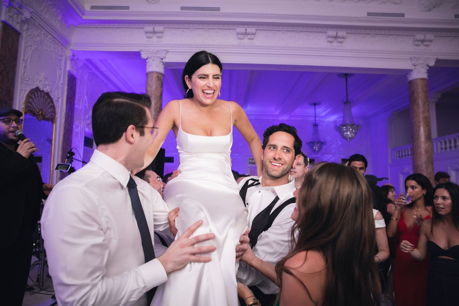 Wedding guests celebrate and dance together at a reception venue with purple ambient lighting.