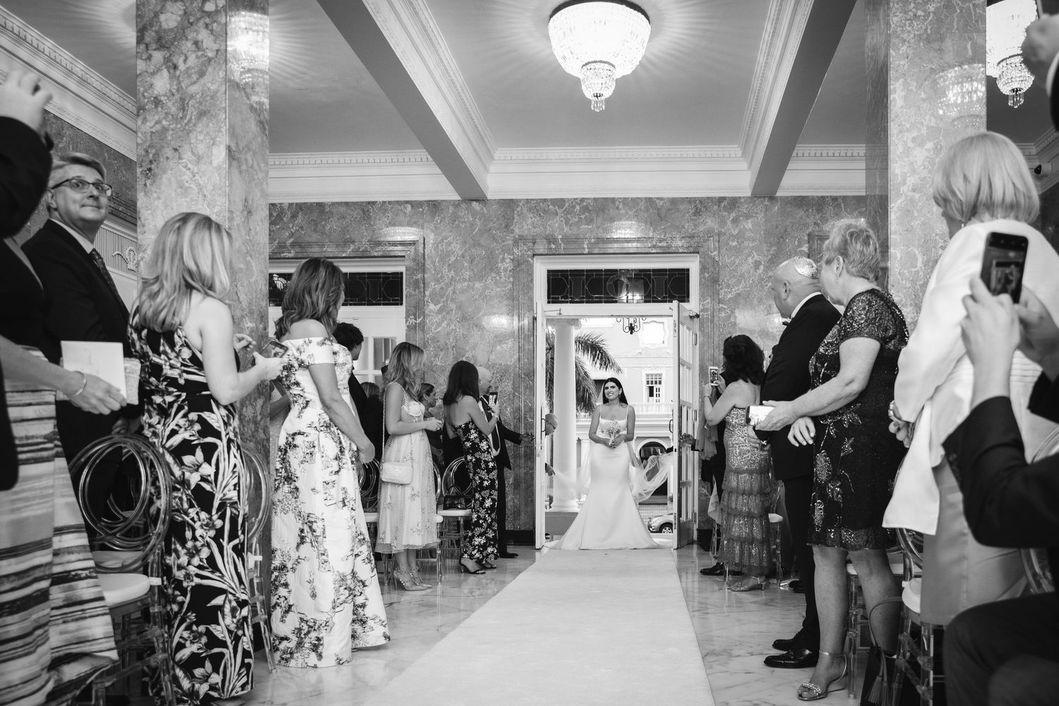 Wedding guests dance and celebrate amid decorative lighting and chandeliers at an elegant indoor reception venue.
