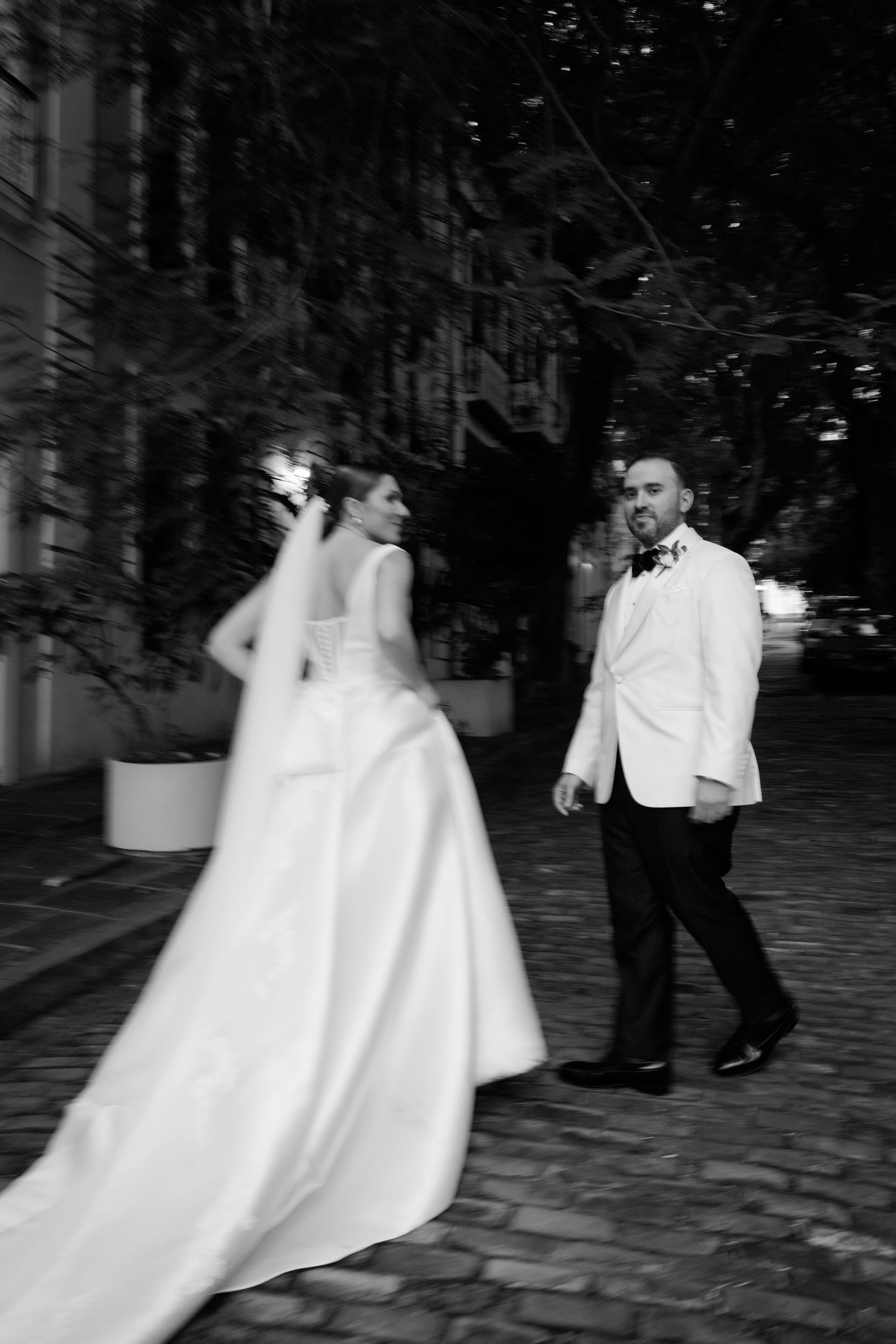 A black and white nighttime photo shows wedding guests dancing and celebrating on a cobblestone street.