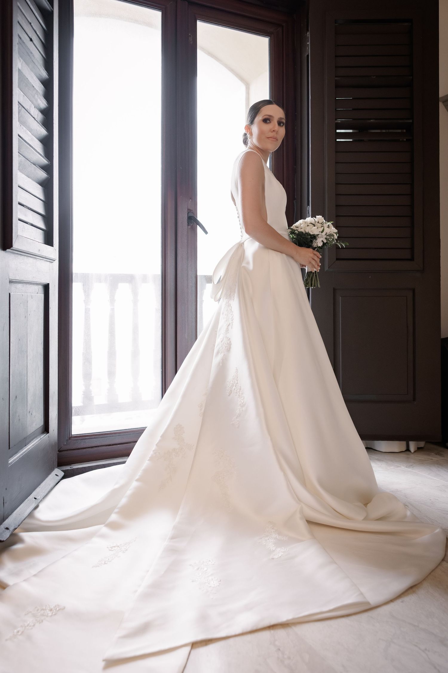 A flowing white wedding dress with a long train elegantly arranged in a hallway with dark shutters and natural light.
