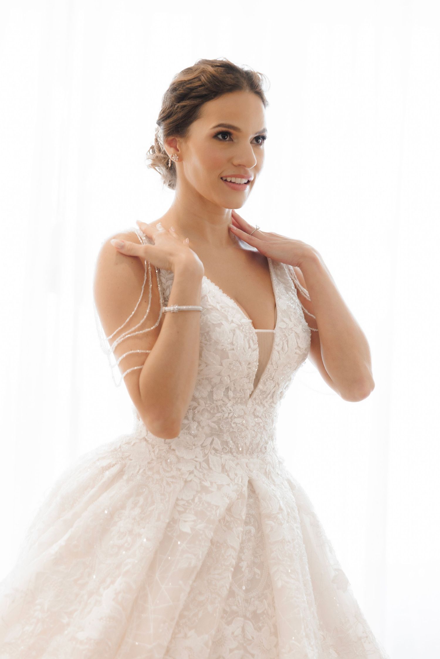 A radiant bride in an elegant white lace ball gown smiles while touching her neck against a bright studio background.