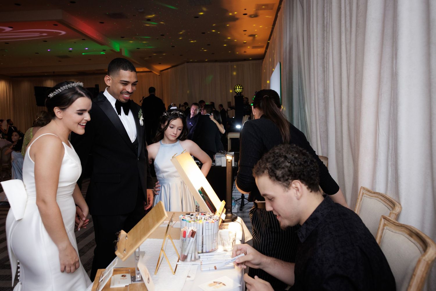 A bride signs the guest book at an elegant wedding reception while guests look on in the background.