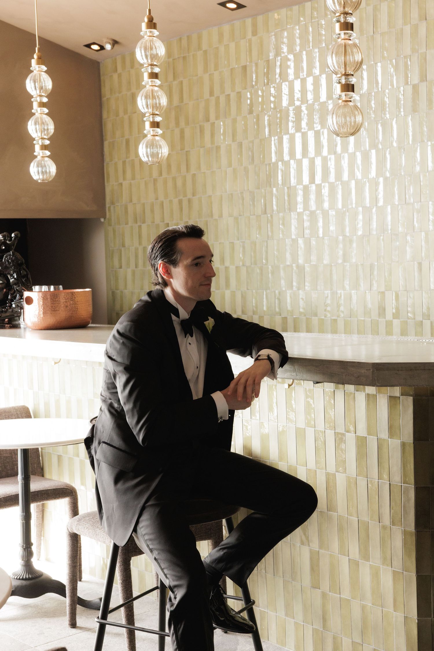 A man in formal attire sits thoughtfully at a modern bar with pendant lighting and textured walls.