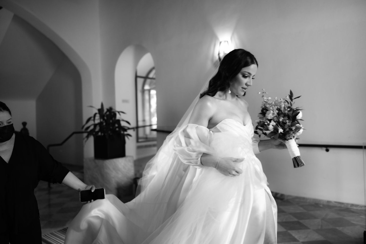 Black and white photo capturing wedding dress preparations in an elegant hallway.