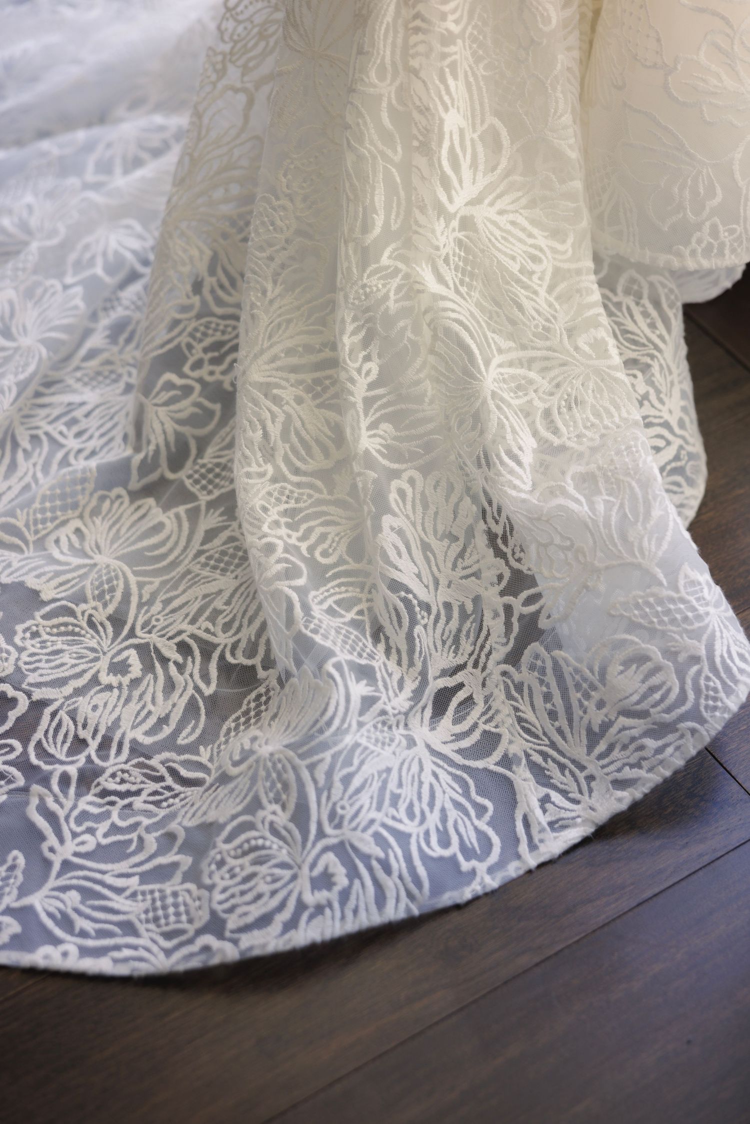 Close-up detail of an intricate lace wedding dress hem against a dark floor.