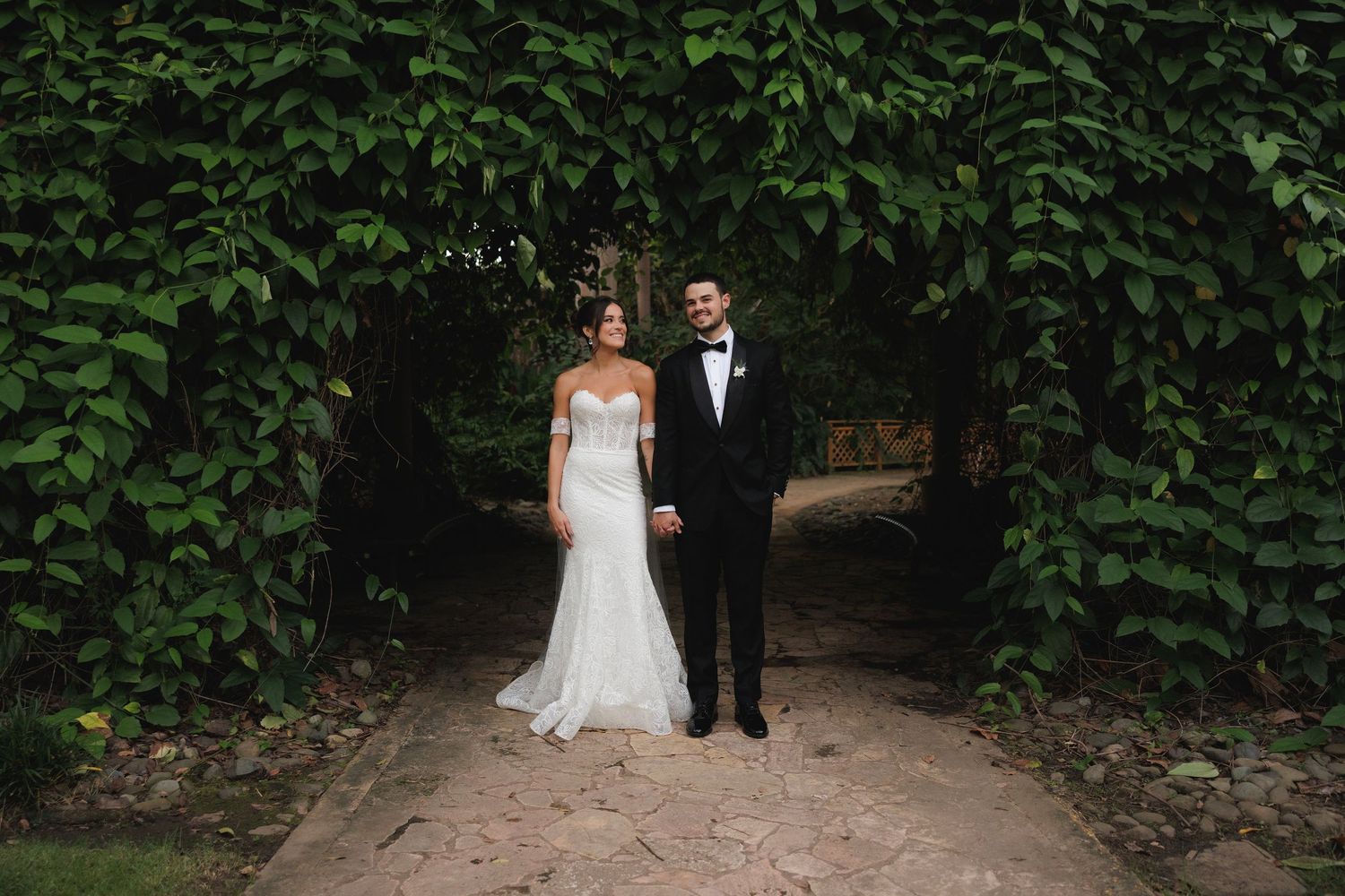 A couple in wedding attire stands together on a garden path surrounded by lush green foliage.