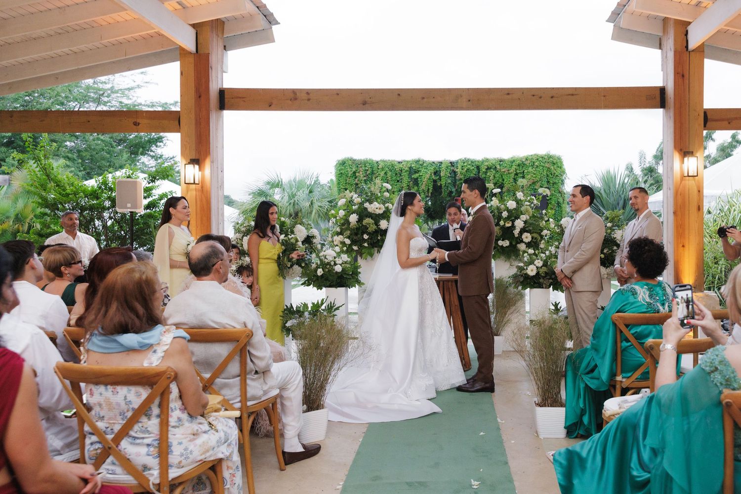 Wedding ceremony takes place under a wooden pergola with guests seated on both sides on a green carpet.