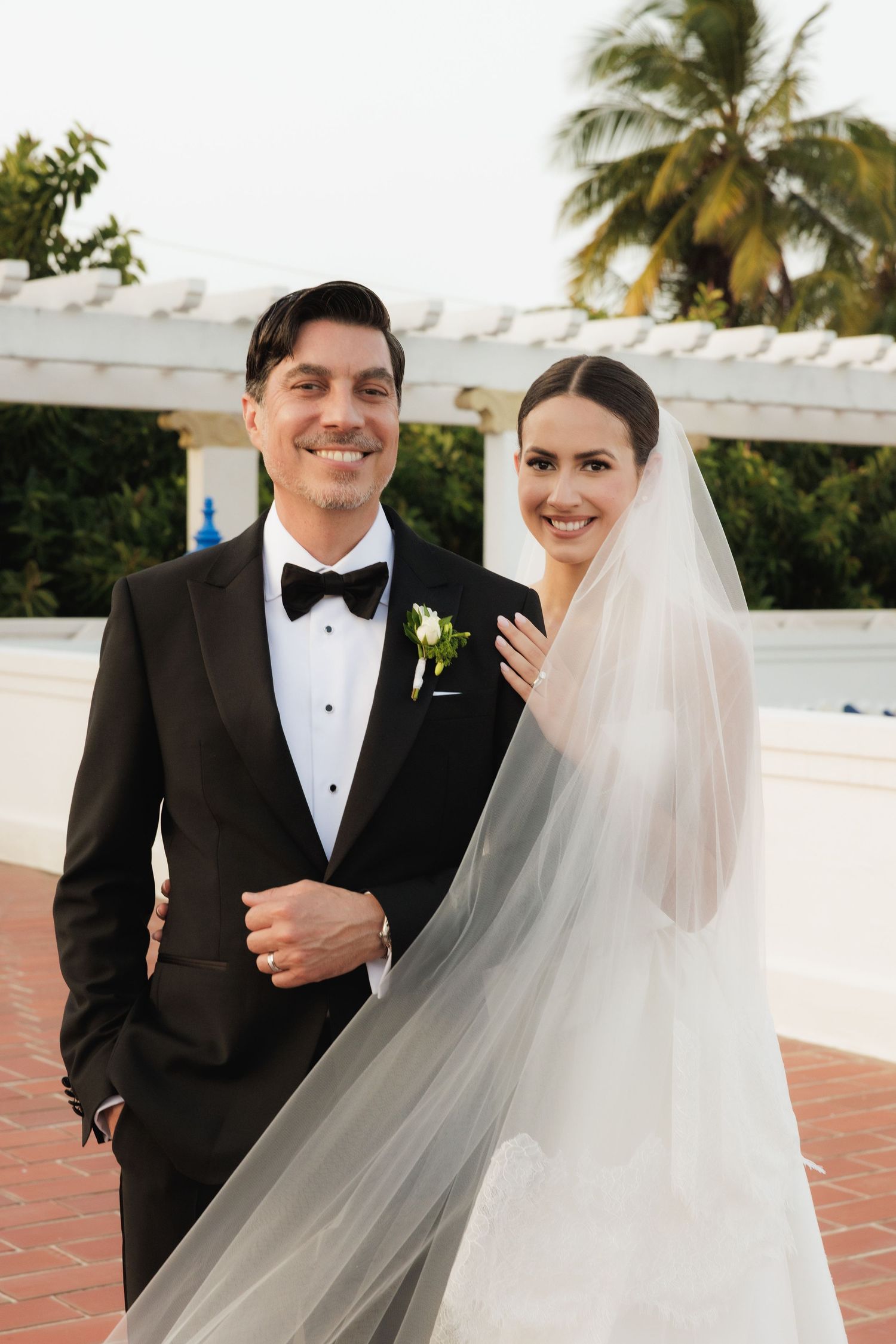 Wedding portrait on rooftop terrace with palm trees and flowing veil in gentle breeze.