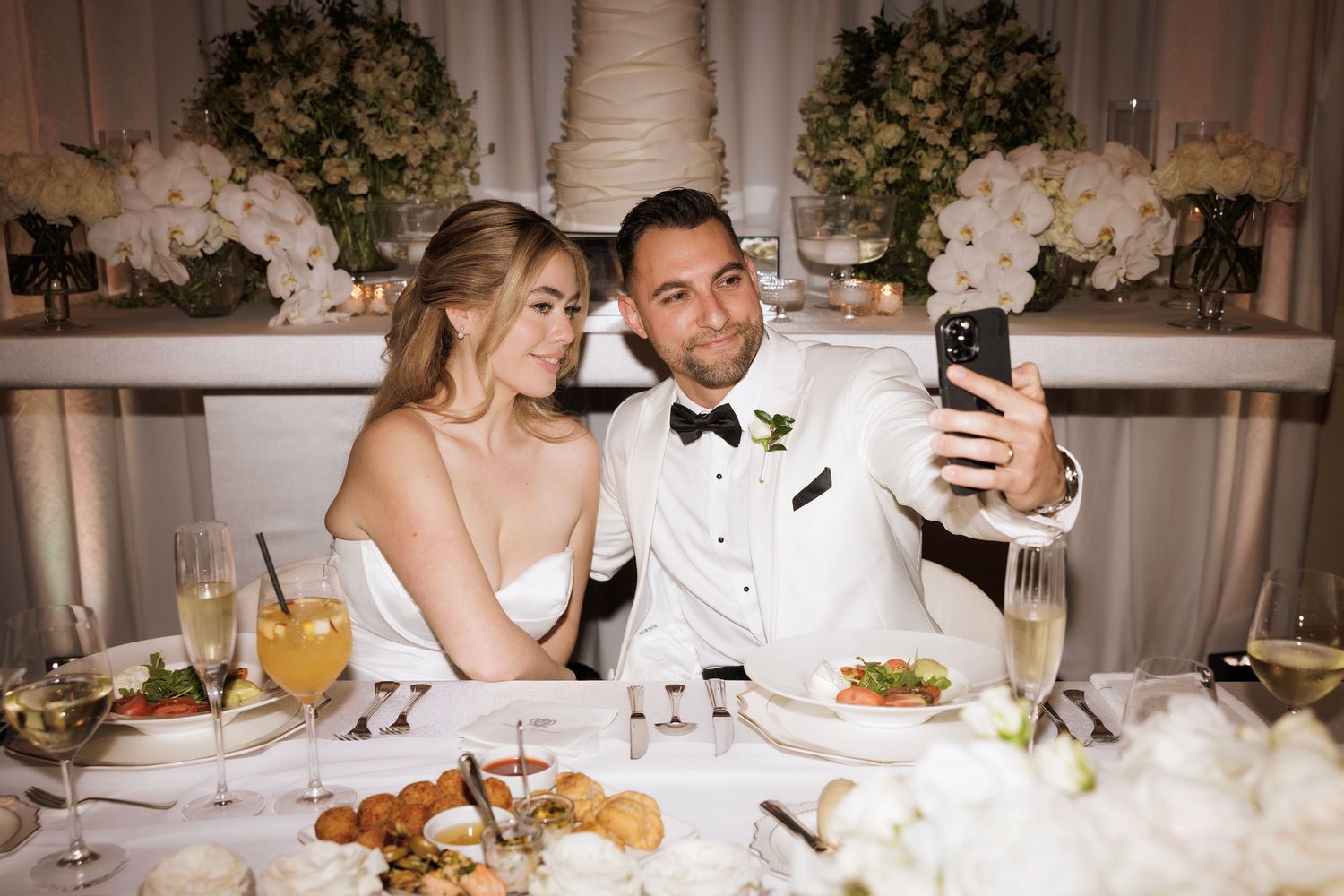 A couple takes a selfie at their wedding reception table decorated with elegant floral arrangements and fine dining settings.