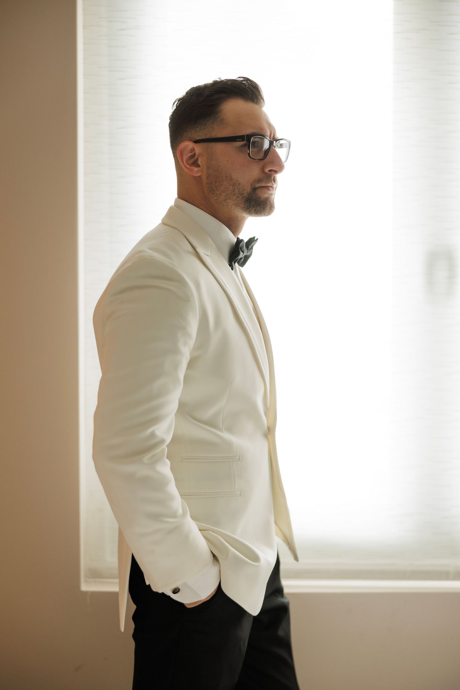 Groom in white tuxedo jacket and bow tie standing by window in classic pose.