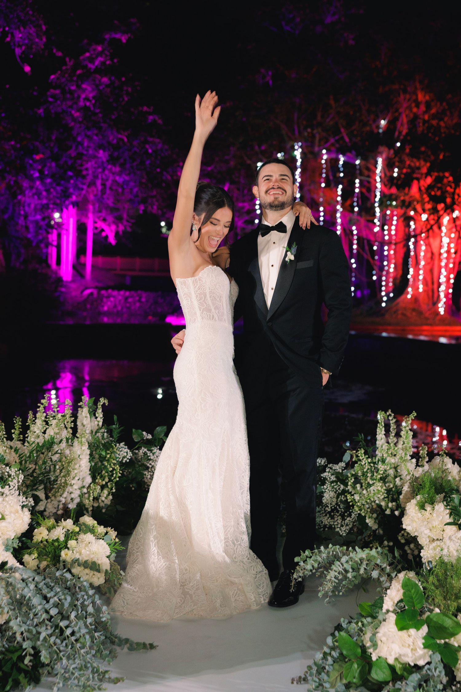 Newlywed couple celebrates on a dance floor surrounded by purple lighting and white floral arrangements at night.