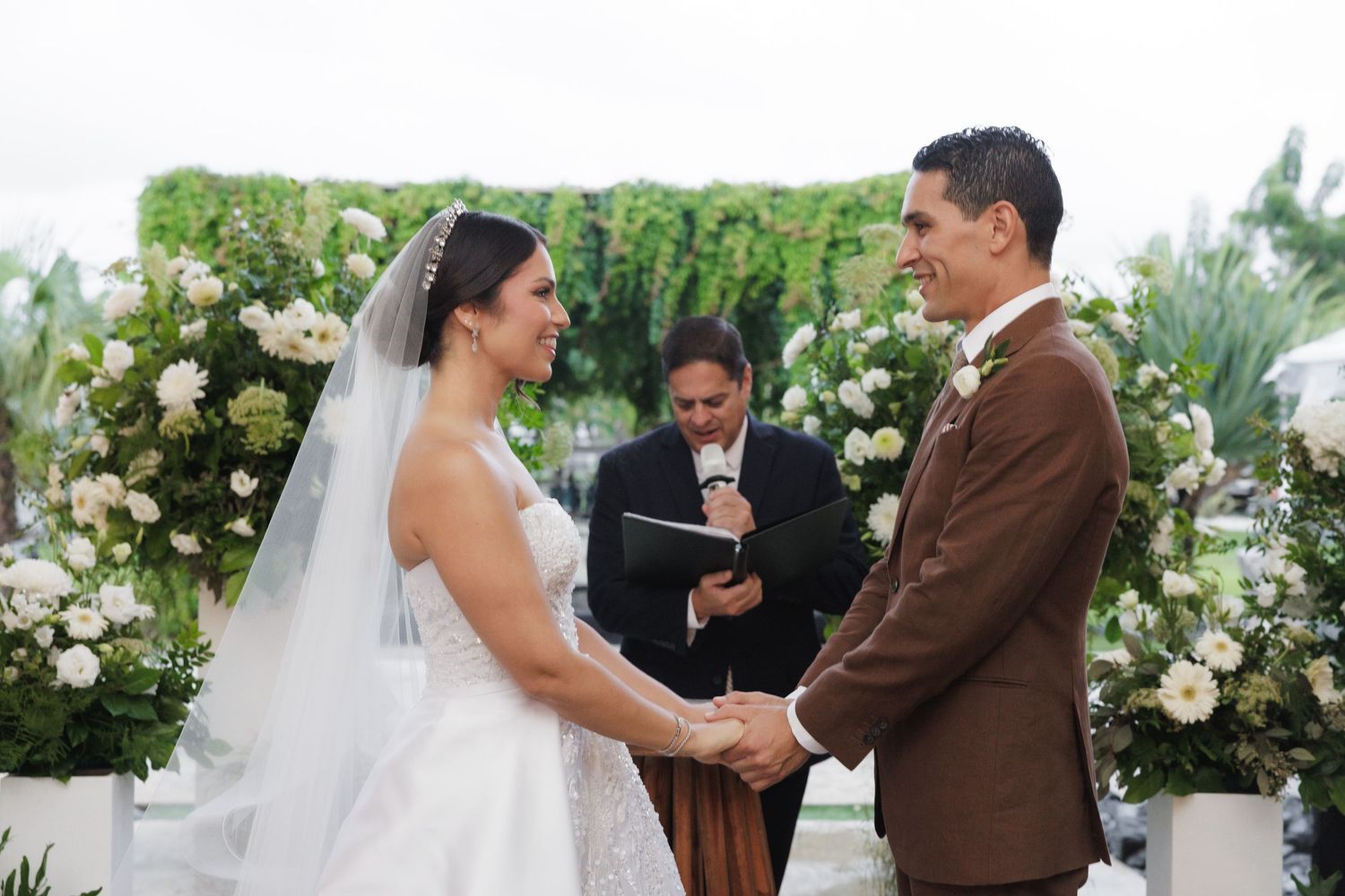 An intimate wedding ceremony takes place under a lush floral arch with white roses and greenery.