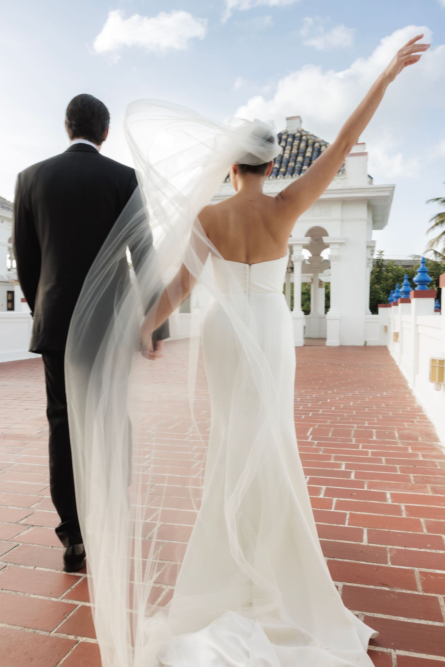 Bride in flowing white gown raises arm joyfully while walking with groom on brick terrace near white pavilion.
