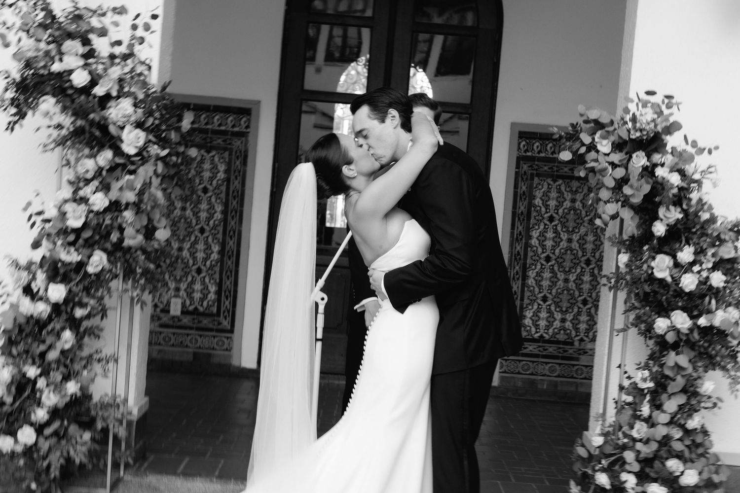 A romantic black and white photograph shows an affectionate moment shared by a couple against a doorway adorned with flowers.
