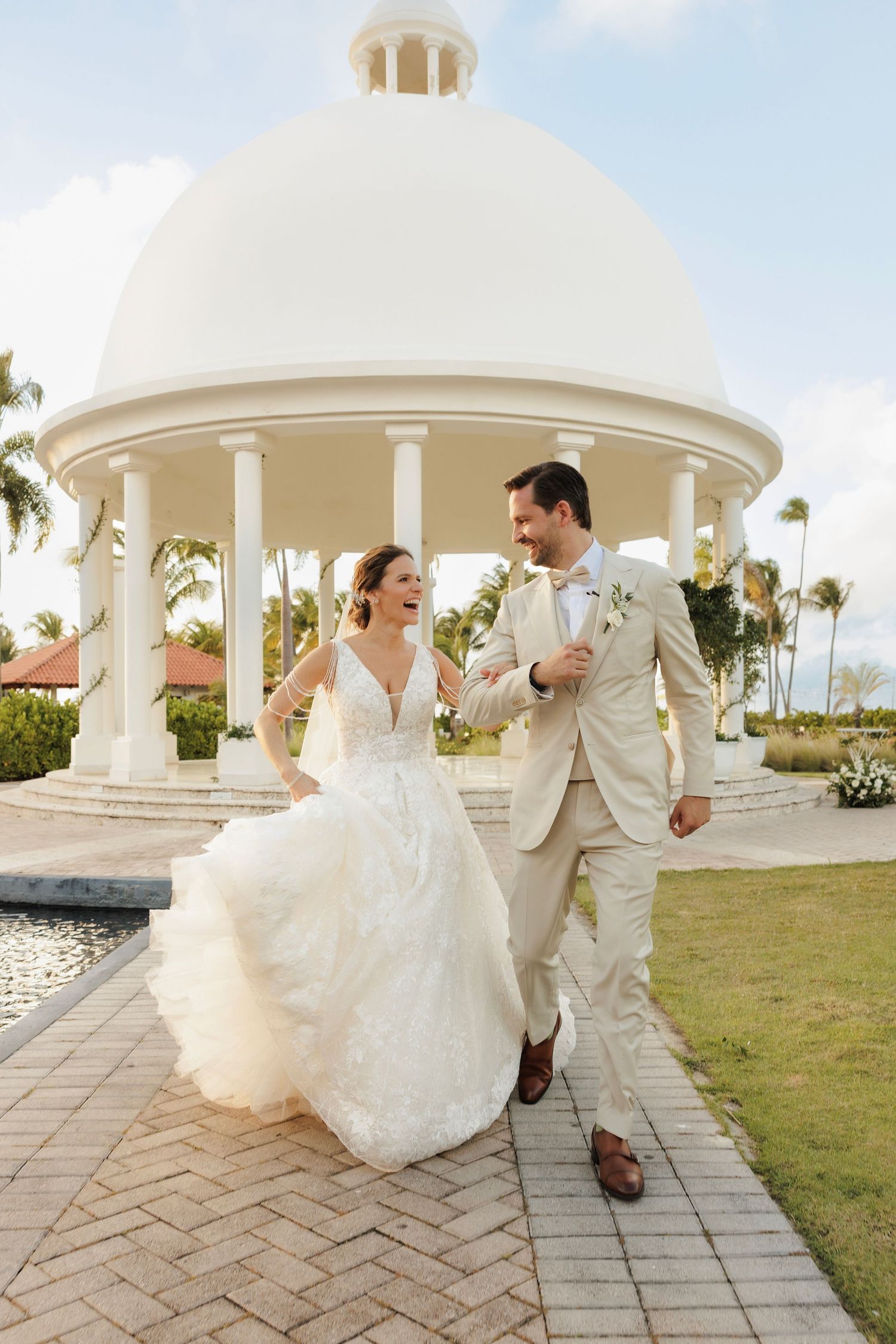 A romantic couple in white attire dances joyfully under an elegant white gazebo by the beach during their special day.