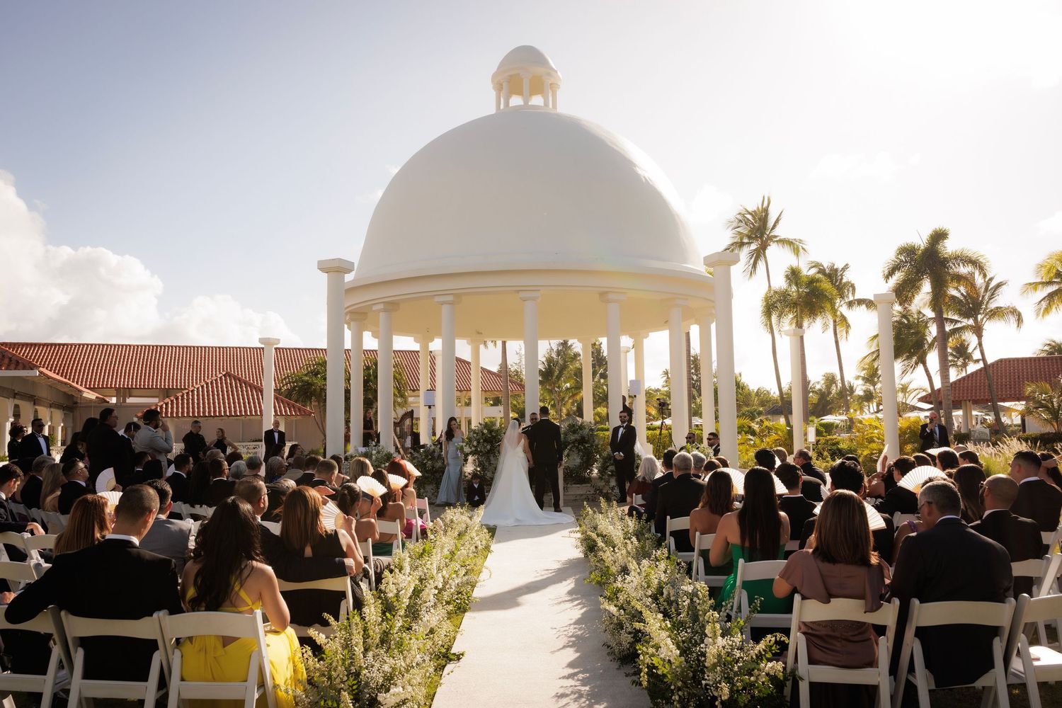 Wedding ceremony setup with white chairs and gazebo overlooking a tropical beach during golden hour.