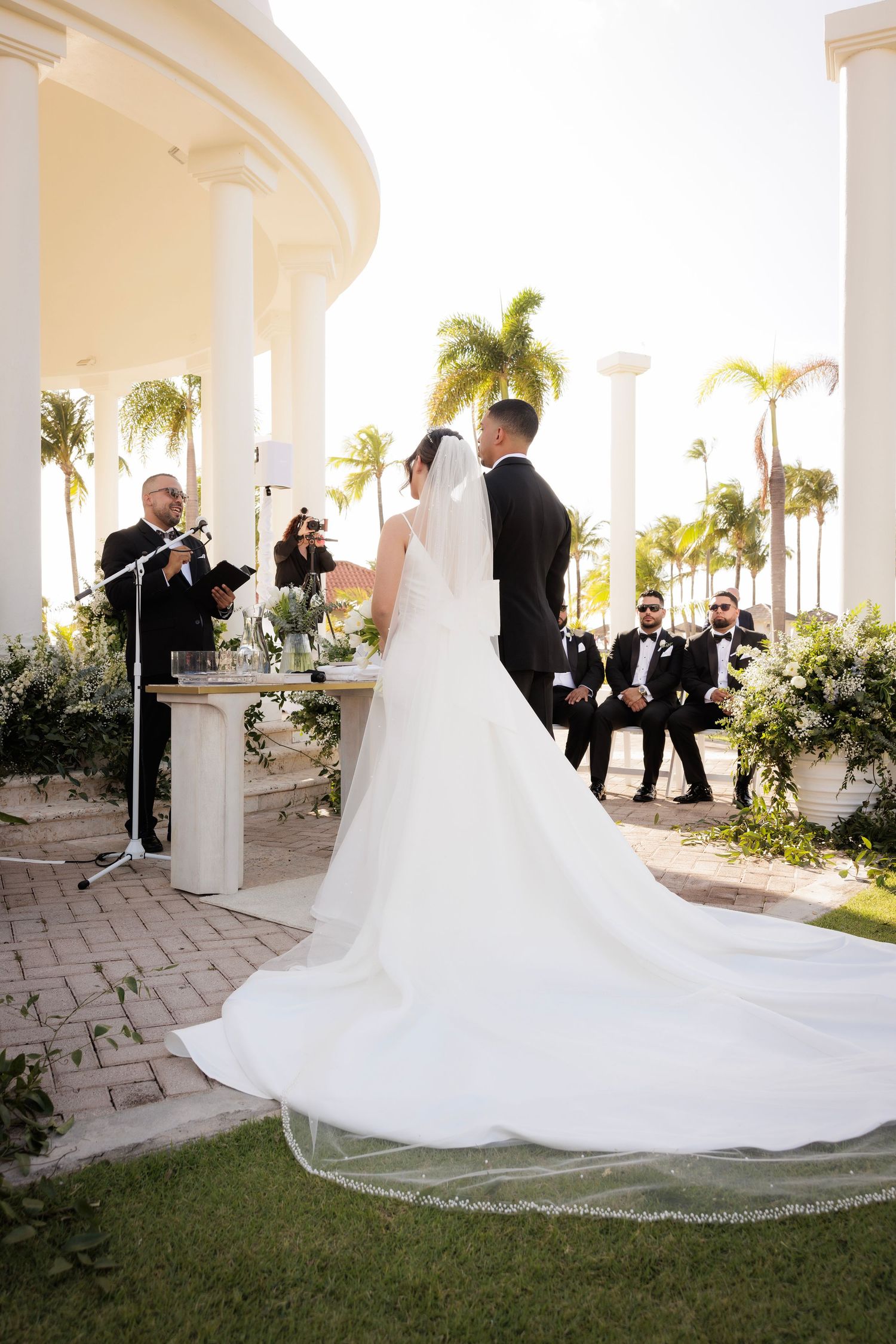 Elegant wedding ceremony on white stone verandah with trailing bridal veil train and swaying palm trees in background.
