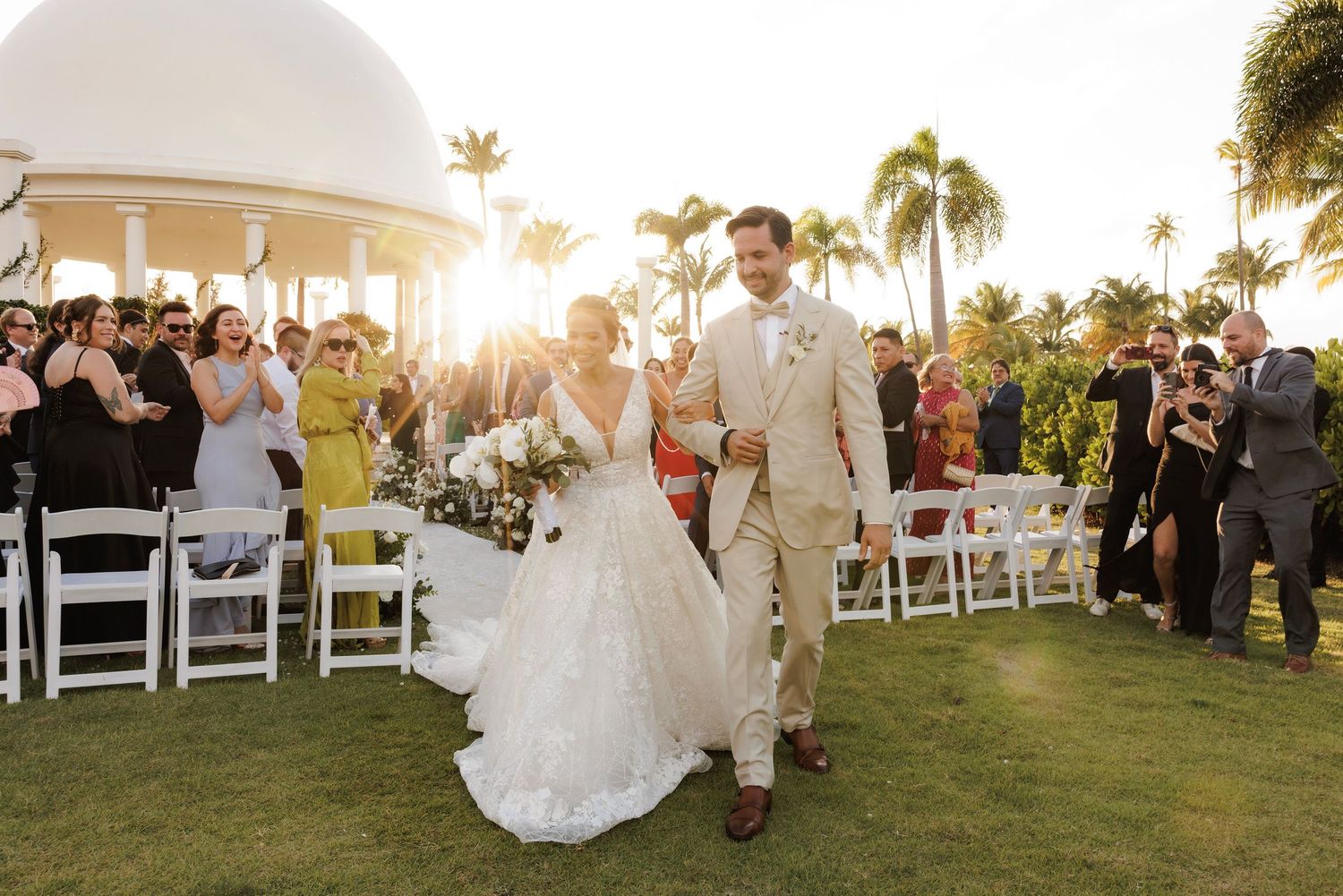 A joyful wedding celebration on a lush green lawn with palm trees and a white gazebo at sunset.