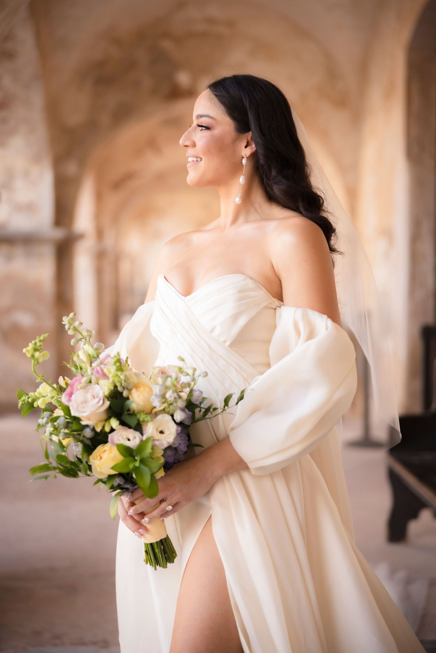 Elegant bride in flowing white dress with puff sleeves holds a spring bouquet in a sunlit stone hallway.