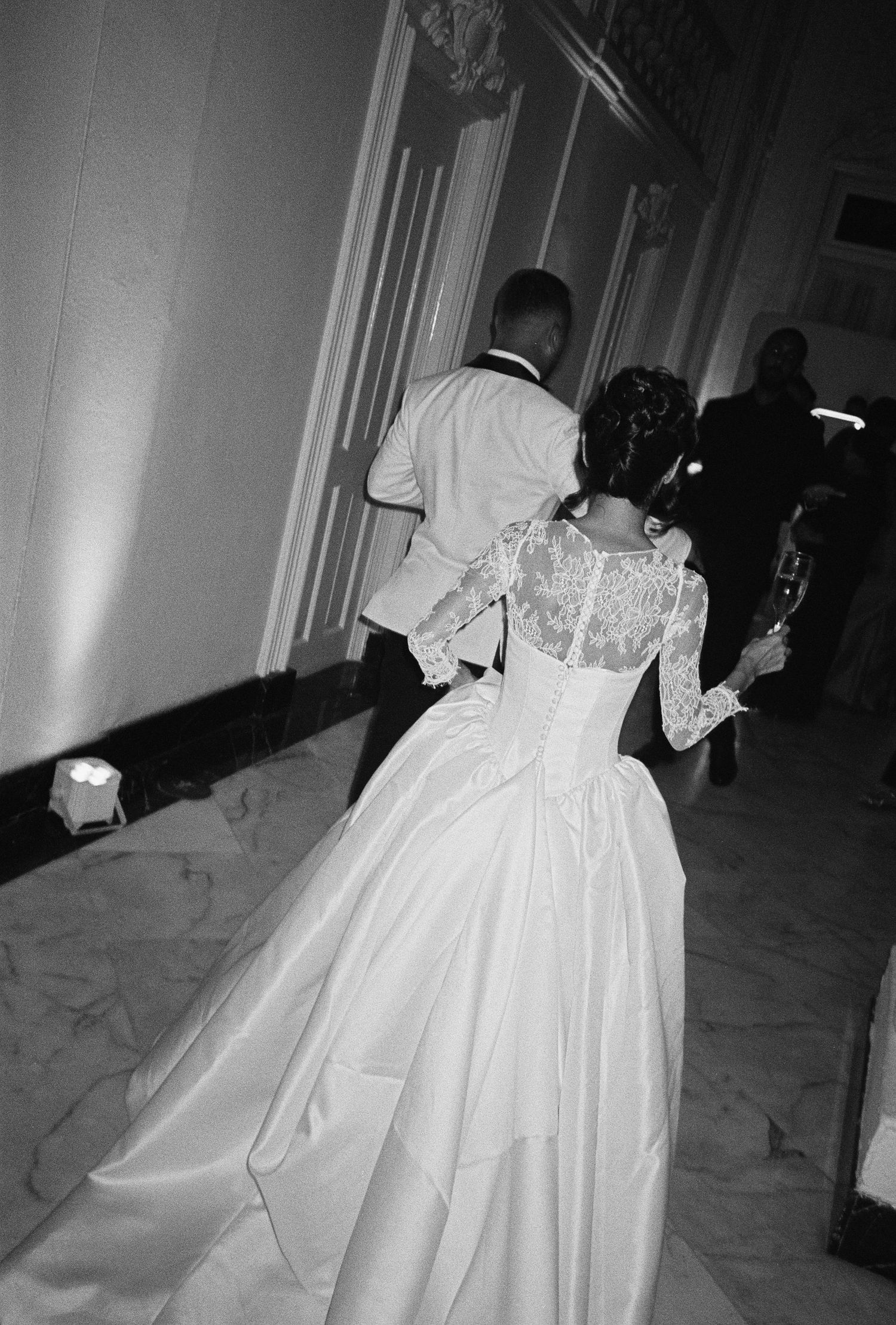 Bride and groom walking in a grand hall, bride wearing a lace back wedding gown with a full skirt.