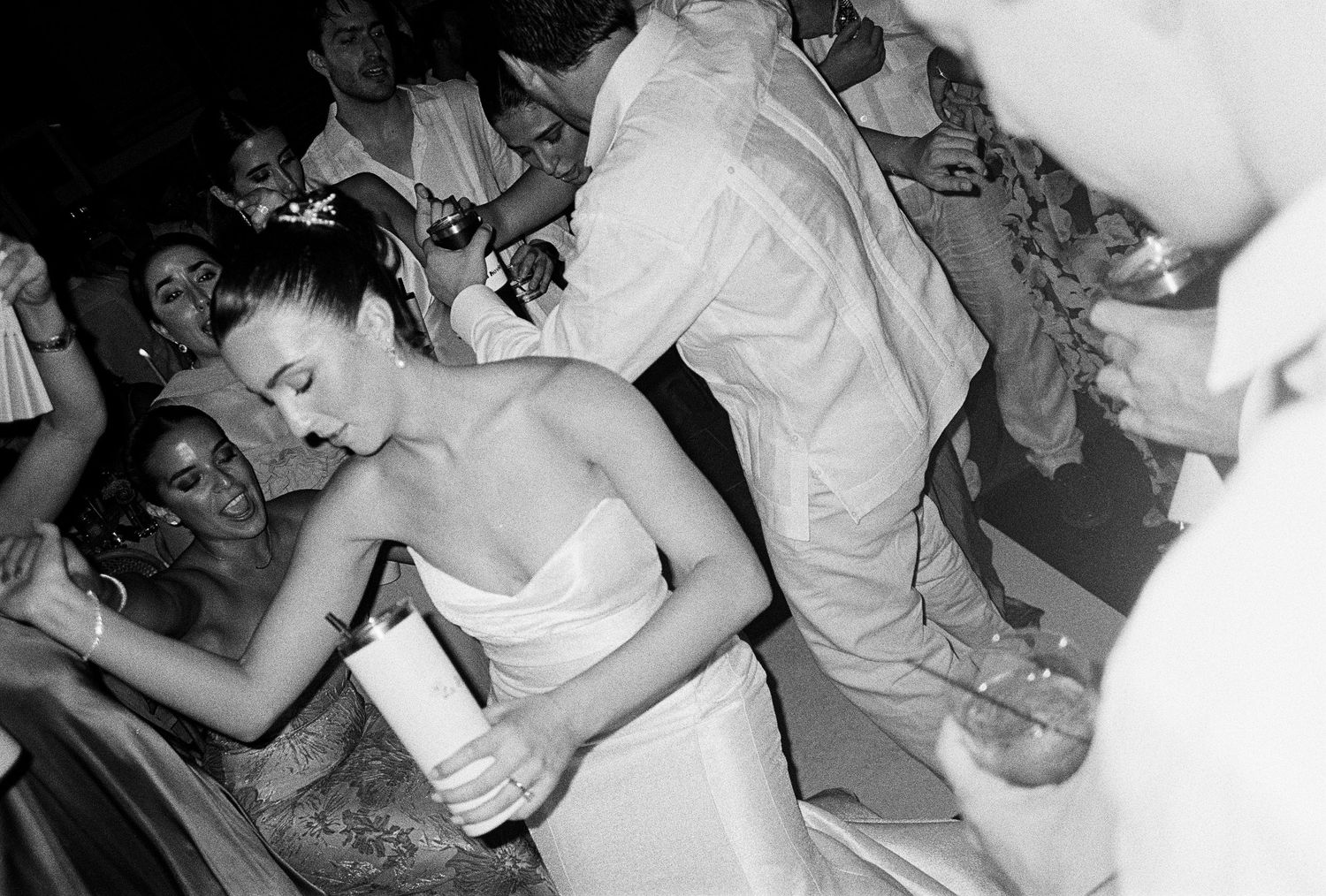 Black and white candid photo of bride in strapless gown mingling with guests at a lively wedding reception.