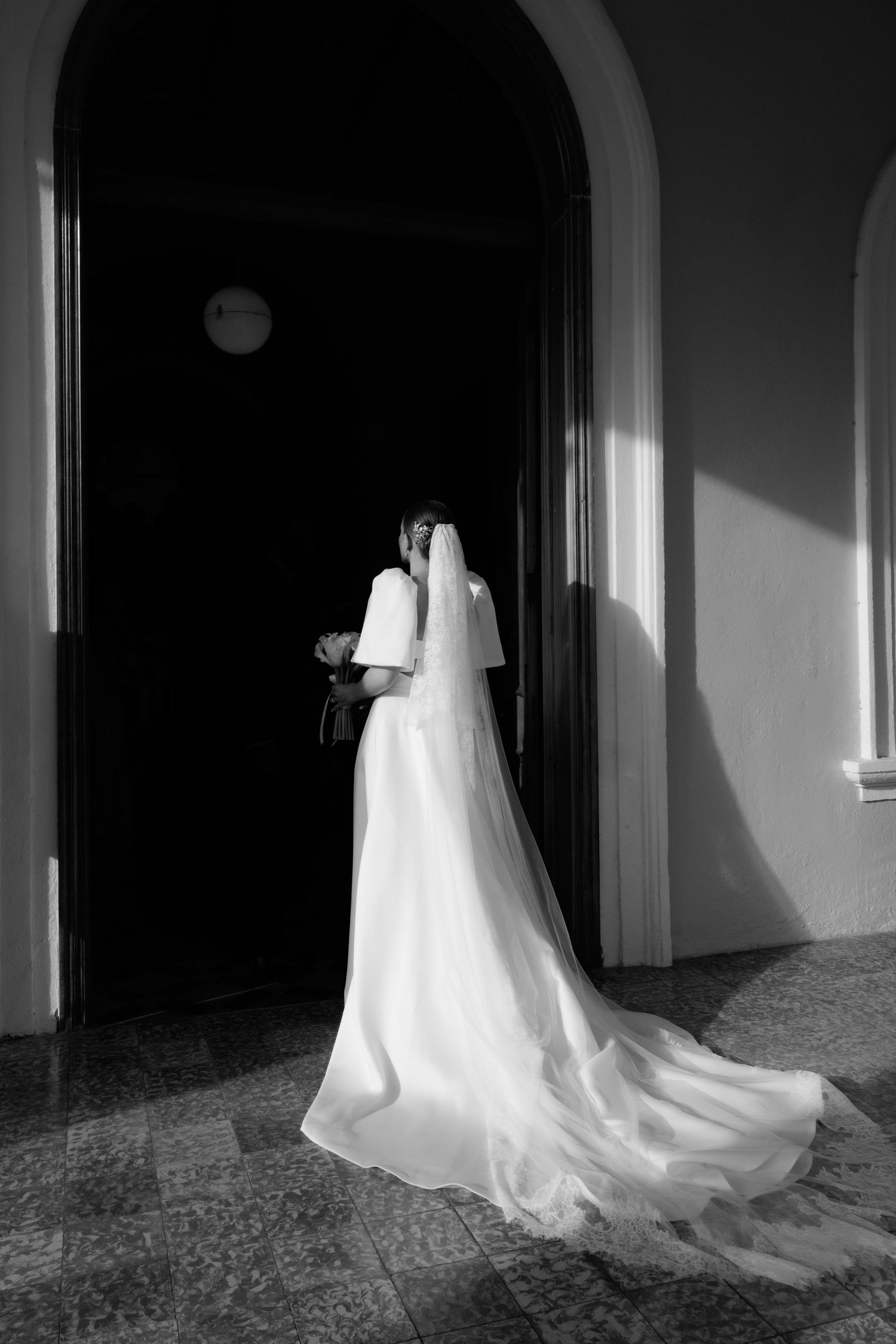 Black and white silhouette of a wedding dress with flowing train in a dramatic archway.