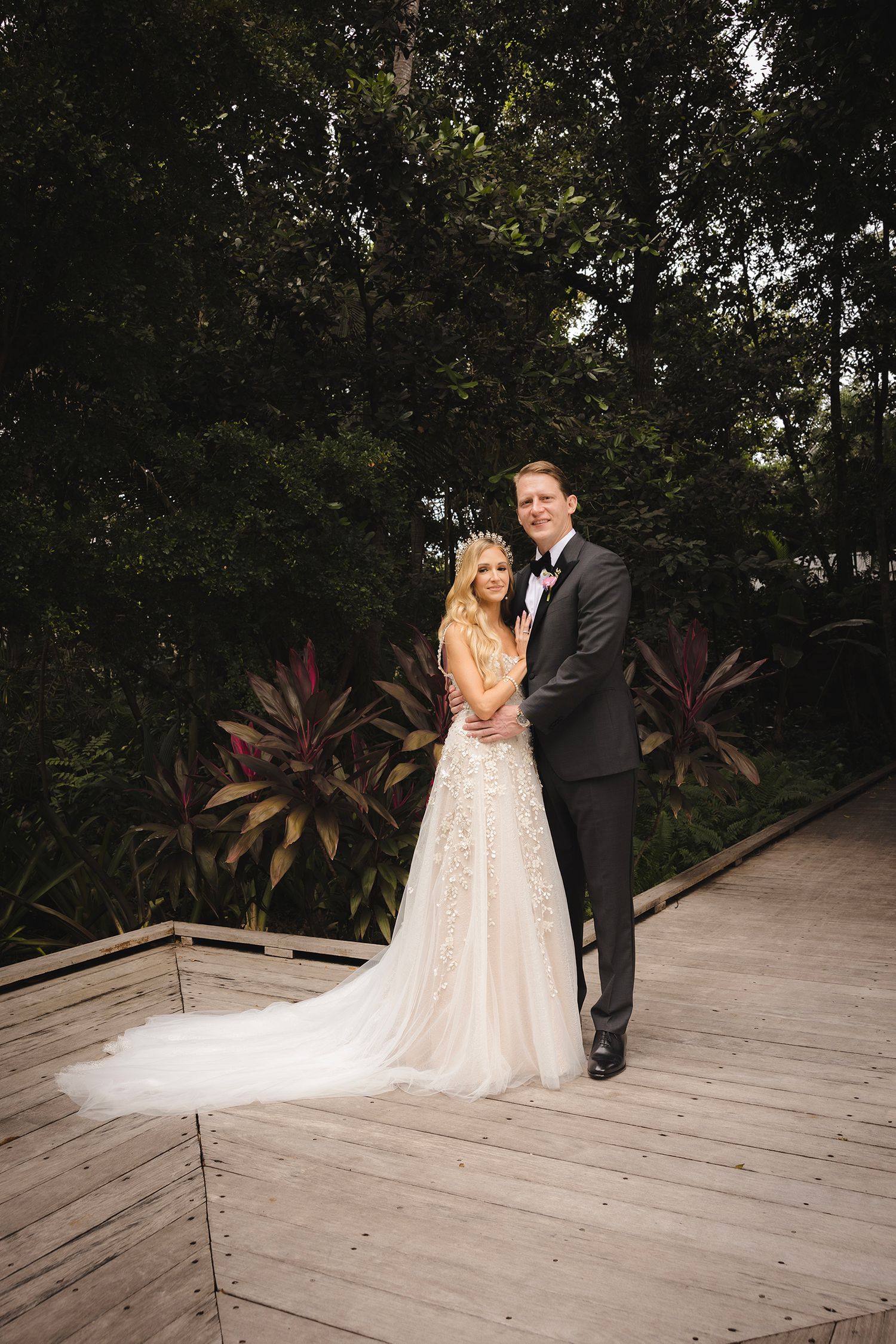 Newlywed couple poses together on wooden deck surrounded by lush tropical plants during outdoor wedding ceremony.