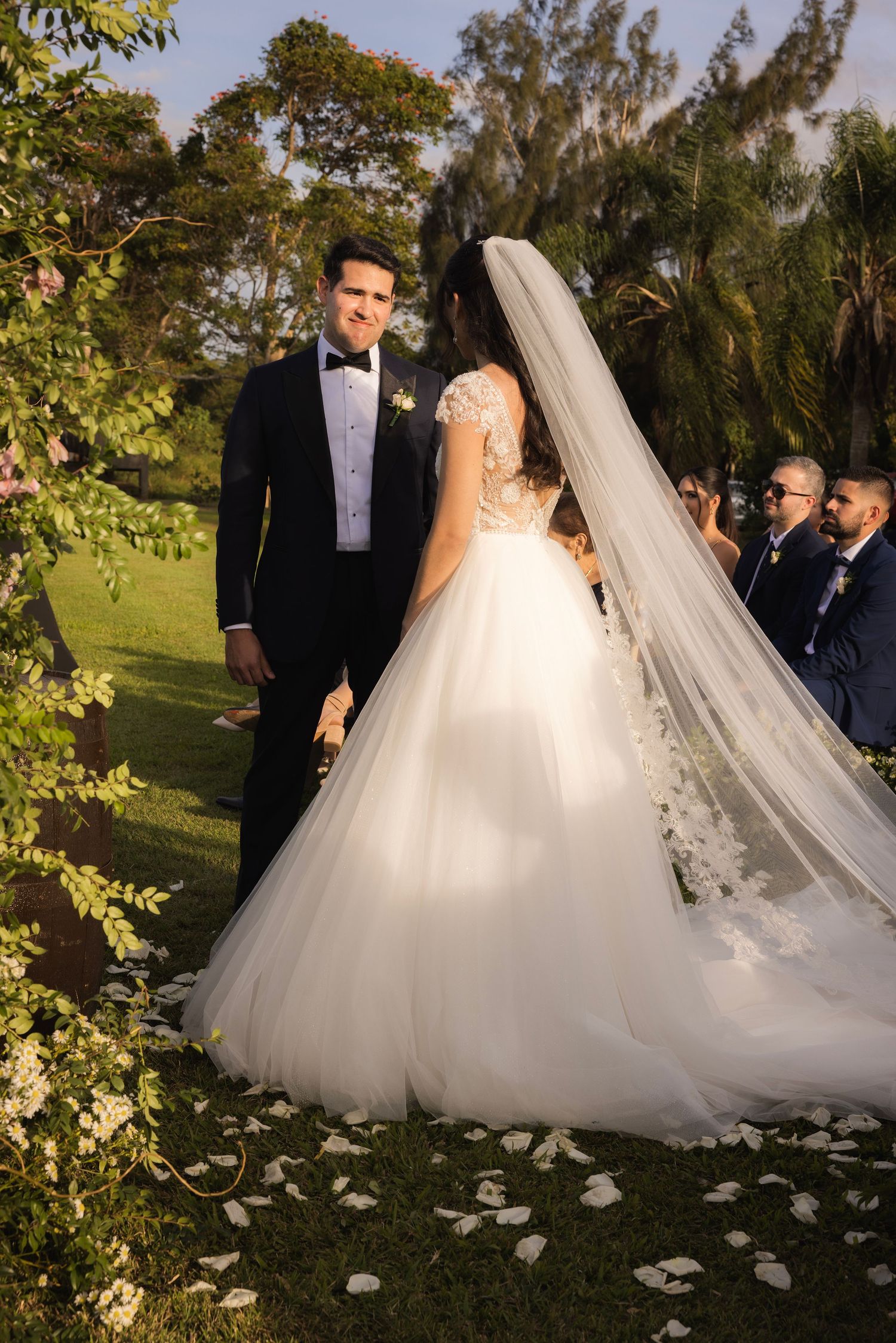 Wedding couple poses in a lush garden setting with a flowing white dress and scattered petals on the ground.