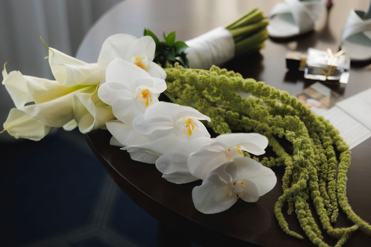 White calla lilies and hanging green amaranth arranged in a dark modern vase on a reflective surface.