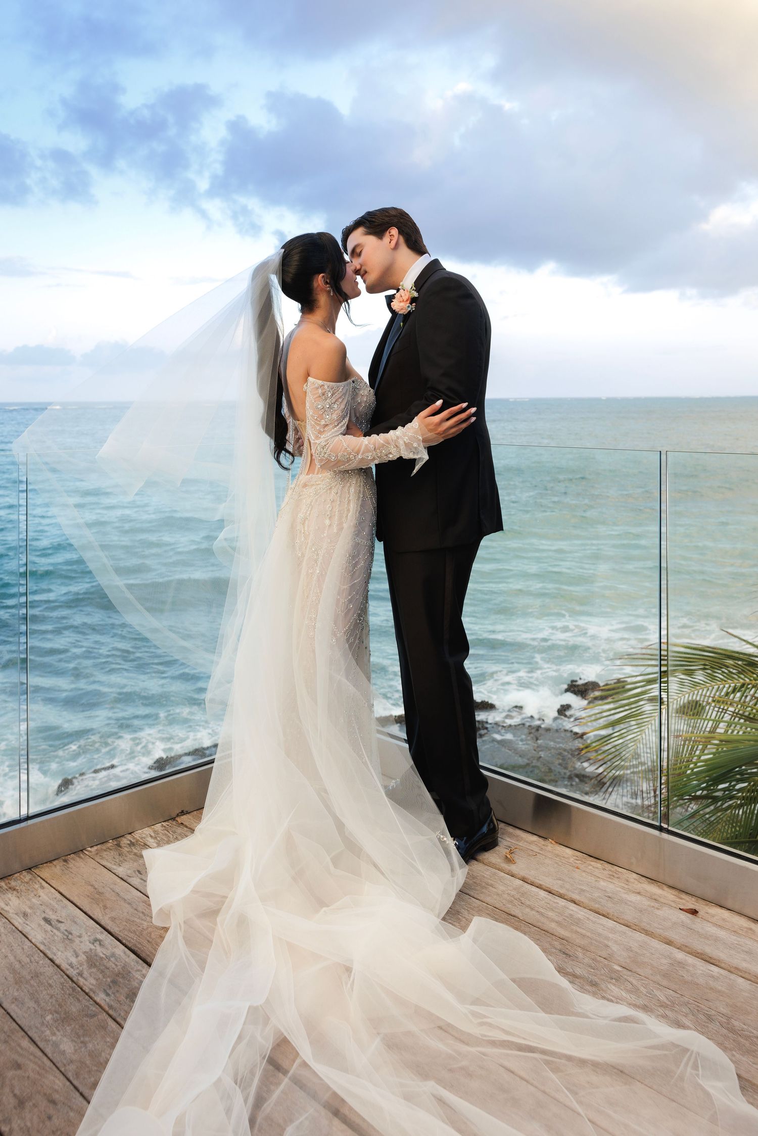 Romantic wedding couple embraces on oceanfront deck with flowing veil and dramatic sky.