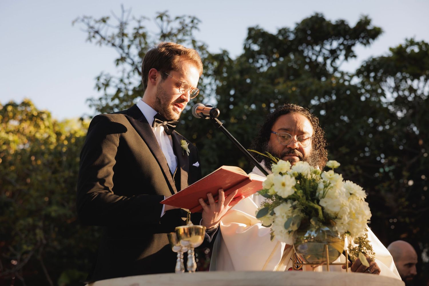 An outdoor wedding ceremony takes place under trees at dusk with a couple exchanging vows before seated guests.