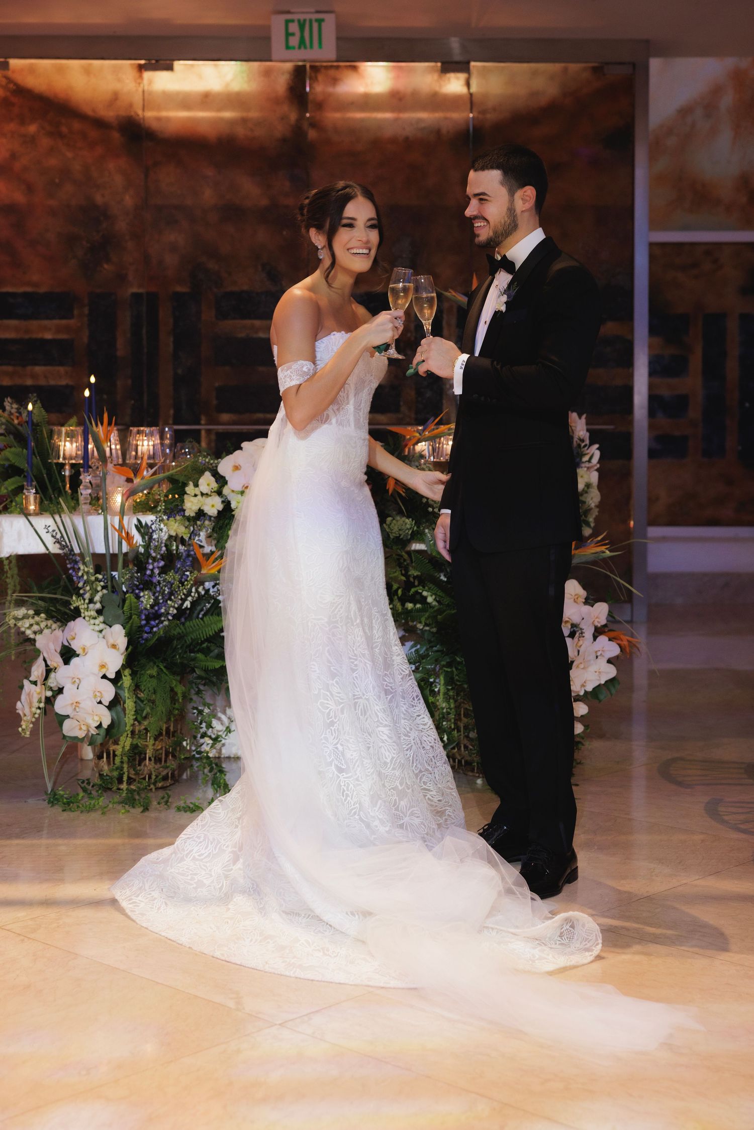Wedding couple toasting champagne at their reception surrounded by elegant floral arrangements and candlelight.