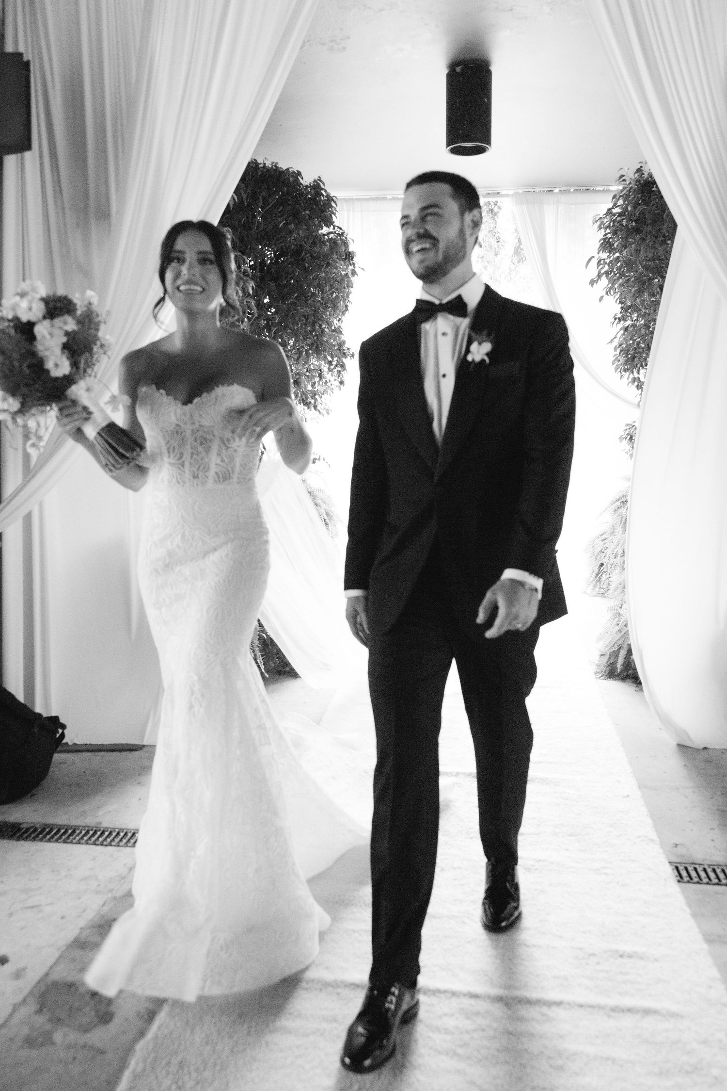 A newlywed couple walks together during their wedding ceremony, with the bride in a fitted white dress and groom in tuxedo.
