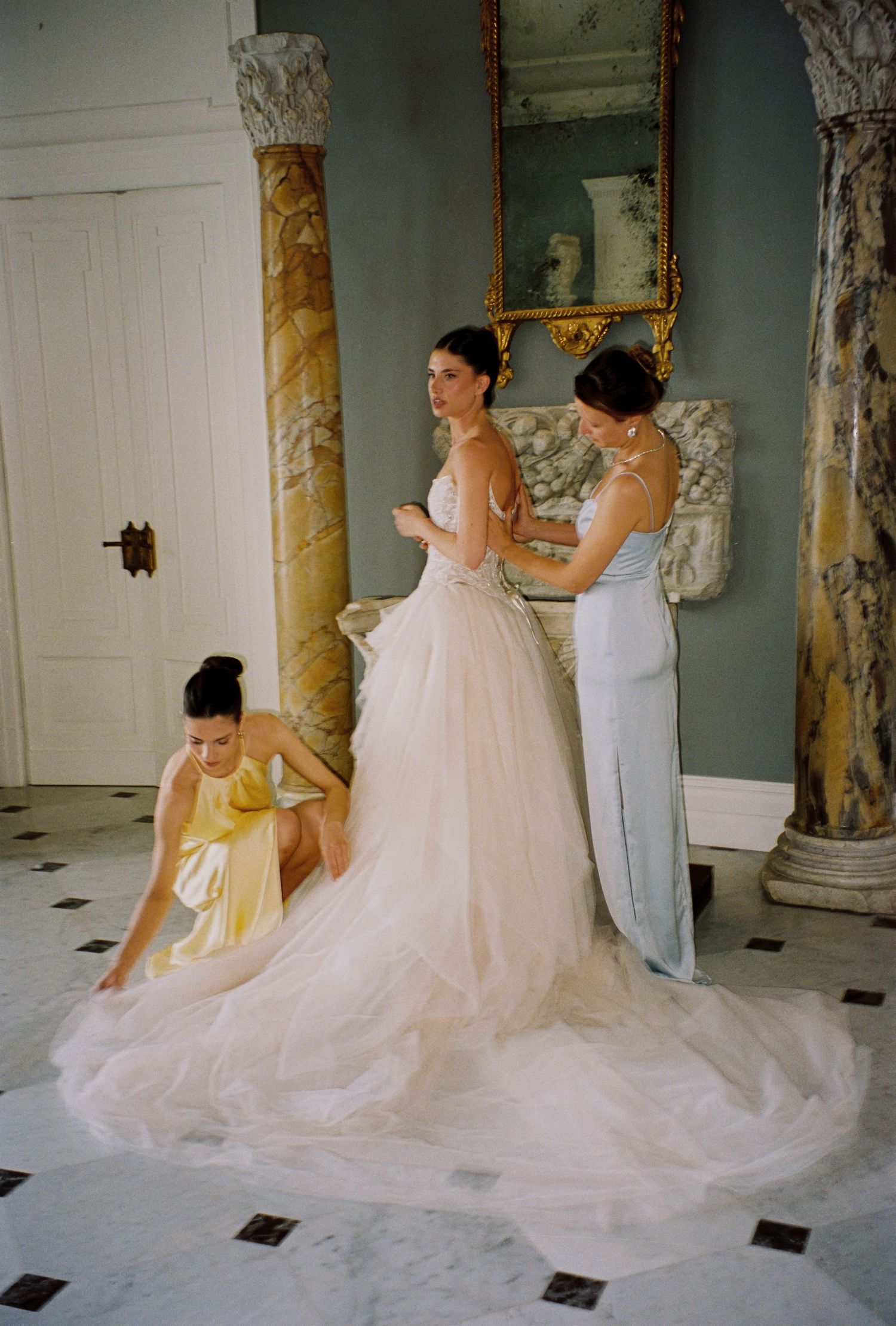 Bride in blush tulle gown assisted by two bridesmaids in elegant room with marble columns and gold mirror.