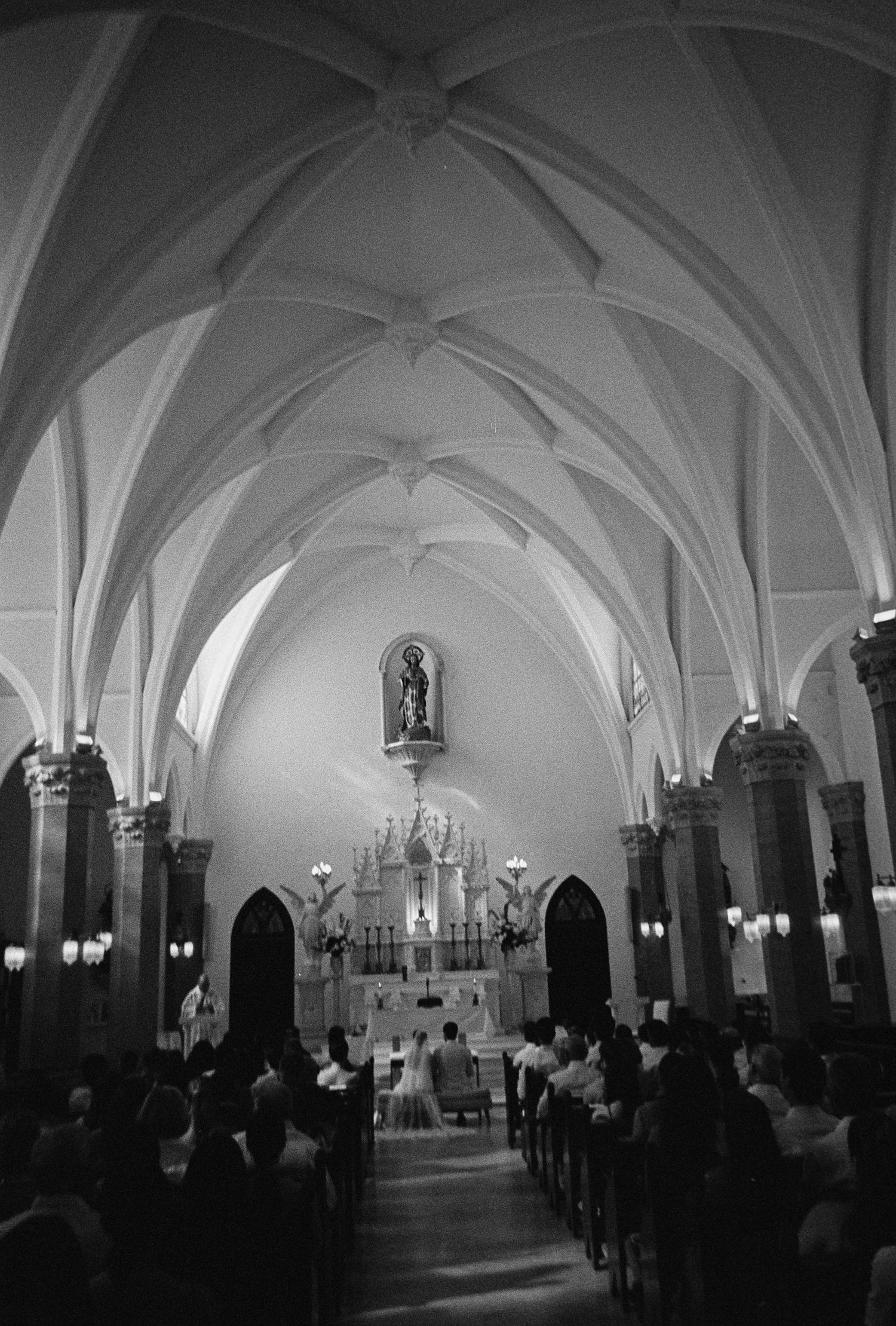 Black and white interior of a Gothic church filled with wedding guests seated in pews during a ceremony.