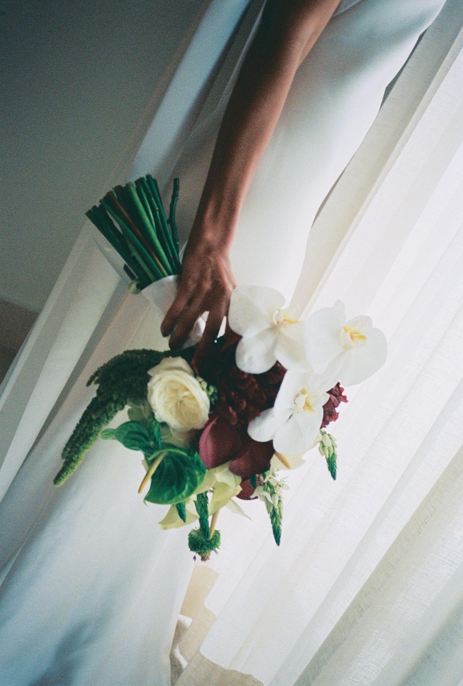 Close-up of bridal bouquet with white orchids and roses held by bride in white dress near window.