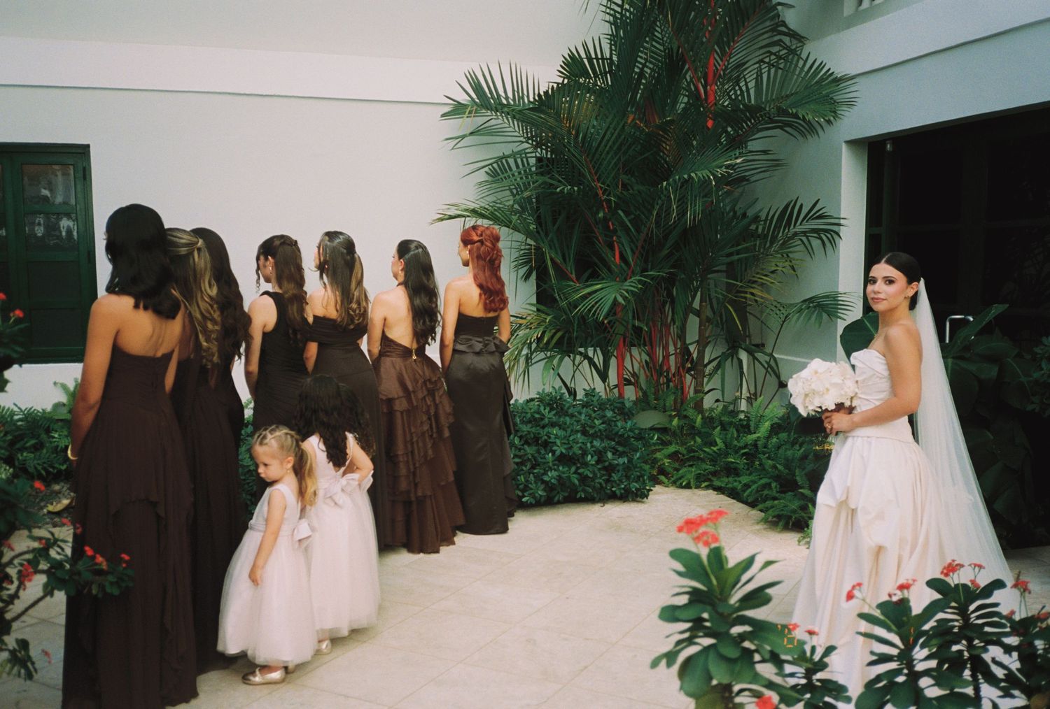 Bride in white gown holds bouquet facing bridesmaids in dark brown dresses with flower girls in pink at outdoor wedding.