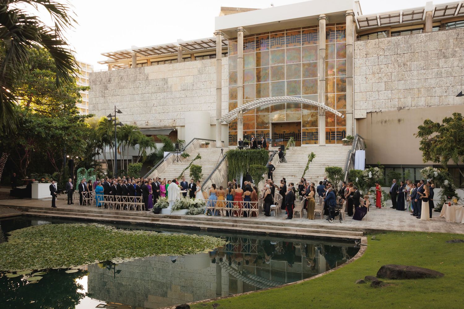 Modern architectural venue with stone walls and glass features surrounded by lush landscaping and outdoor gathering spaces.