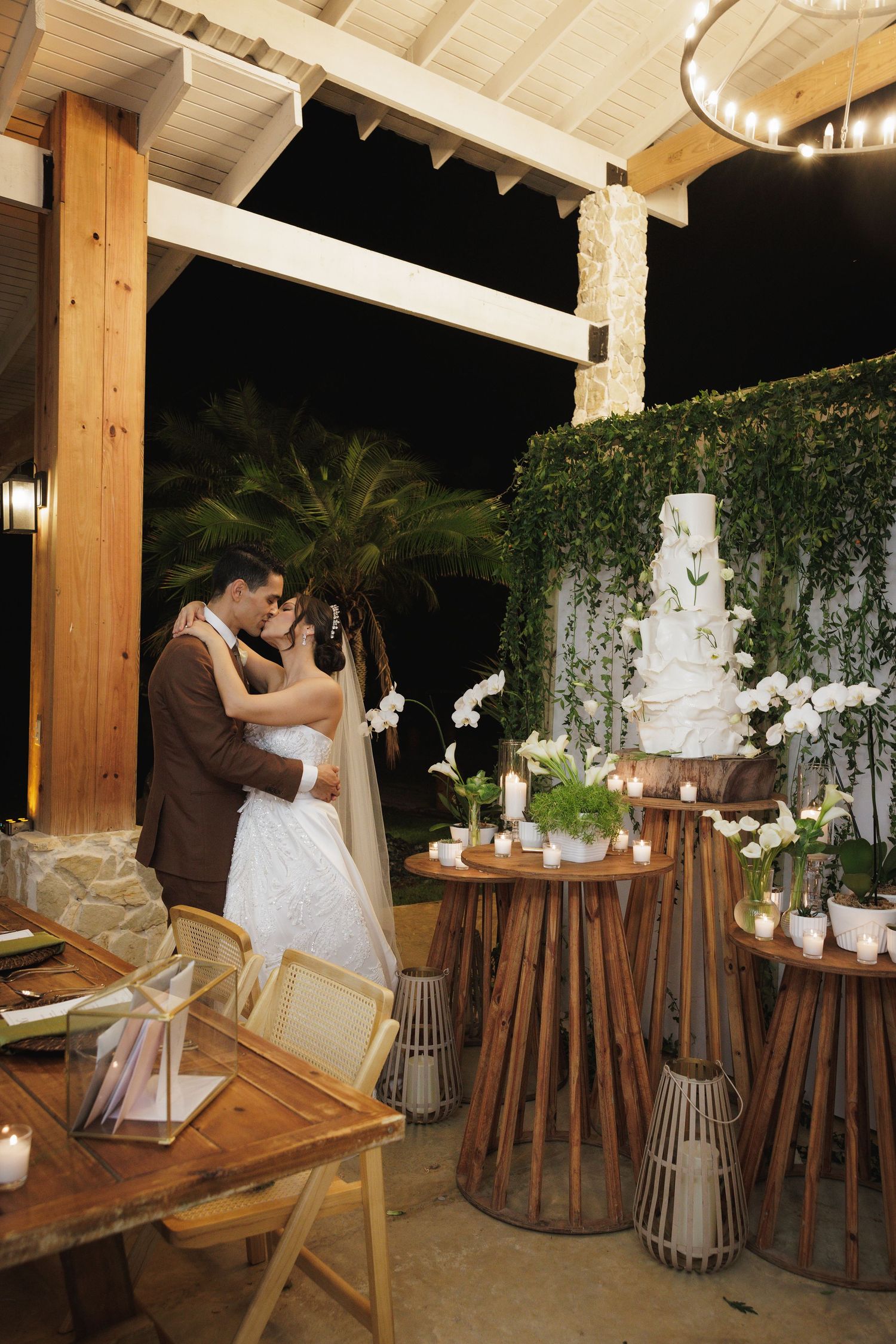 Romantic outdoor wedding reception setup with wooden decor, white floral arrangements, and warm lighting under a covered patio.