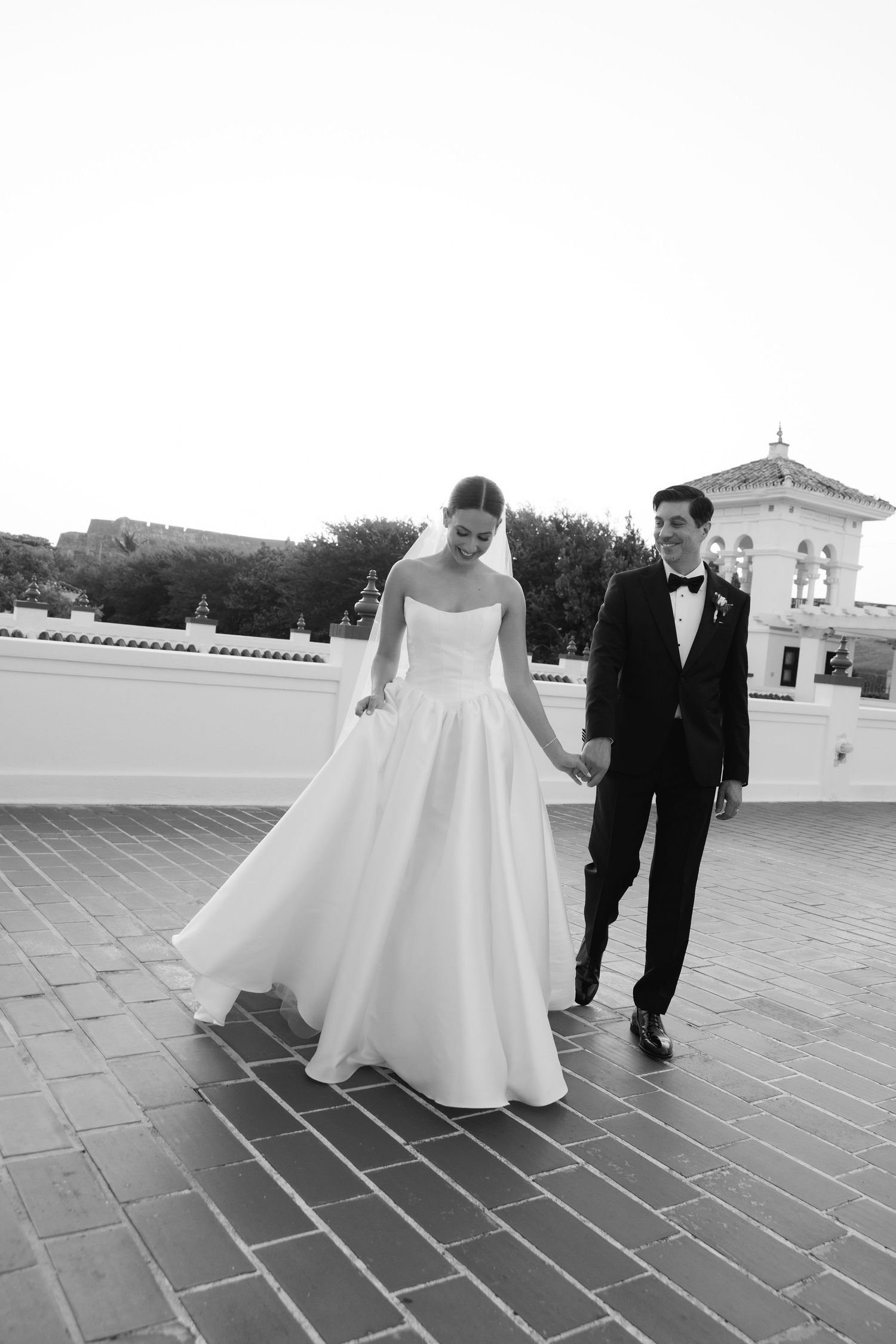 Black and white photo of newlyweds walking together on rooftop with architectural details.