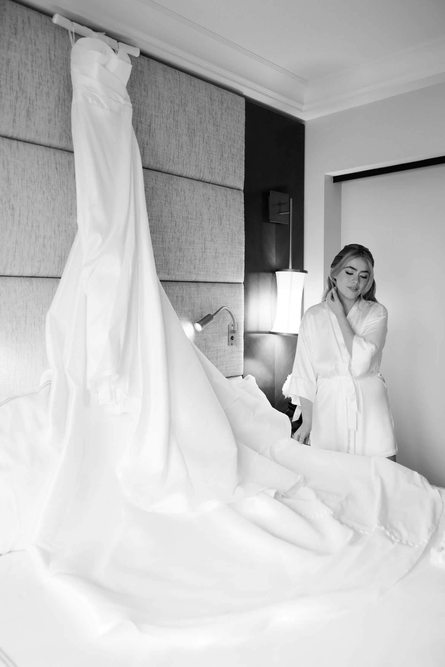 White wedding dress hanging in hotel room with natural light streaming through windows.