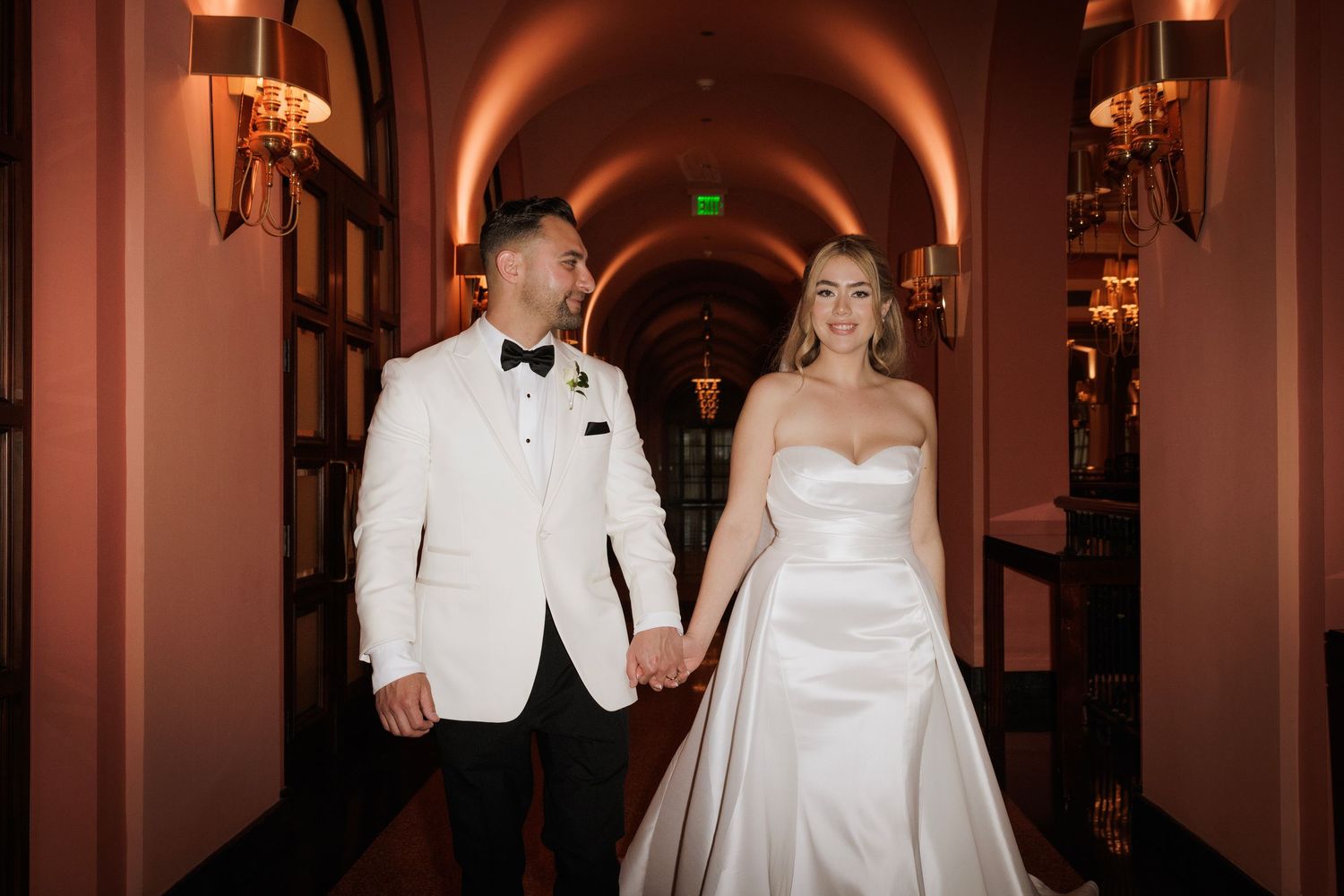 A wedding couple shares a romantic moment in an elegantly lit hallway with warm amber lighting.