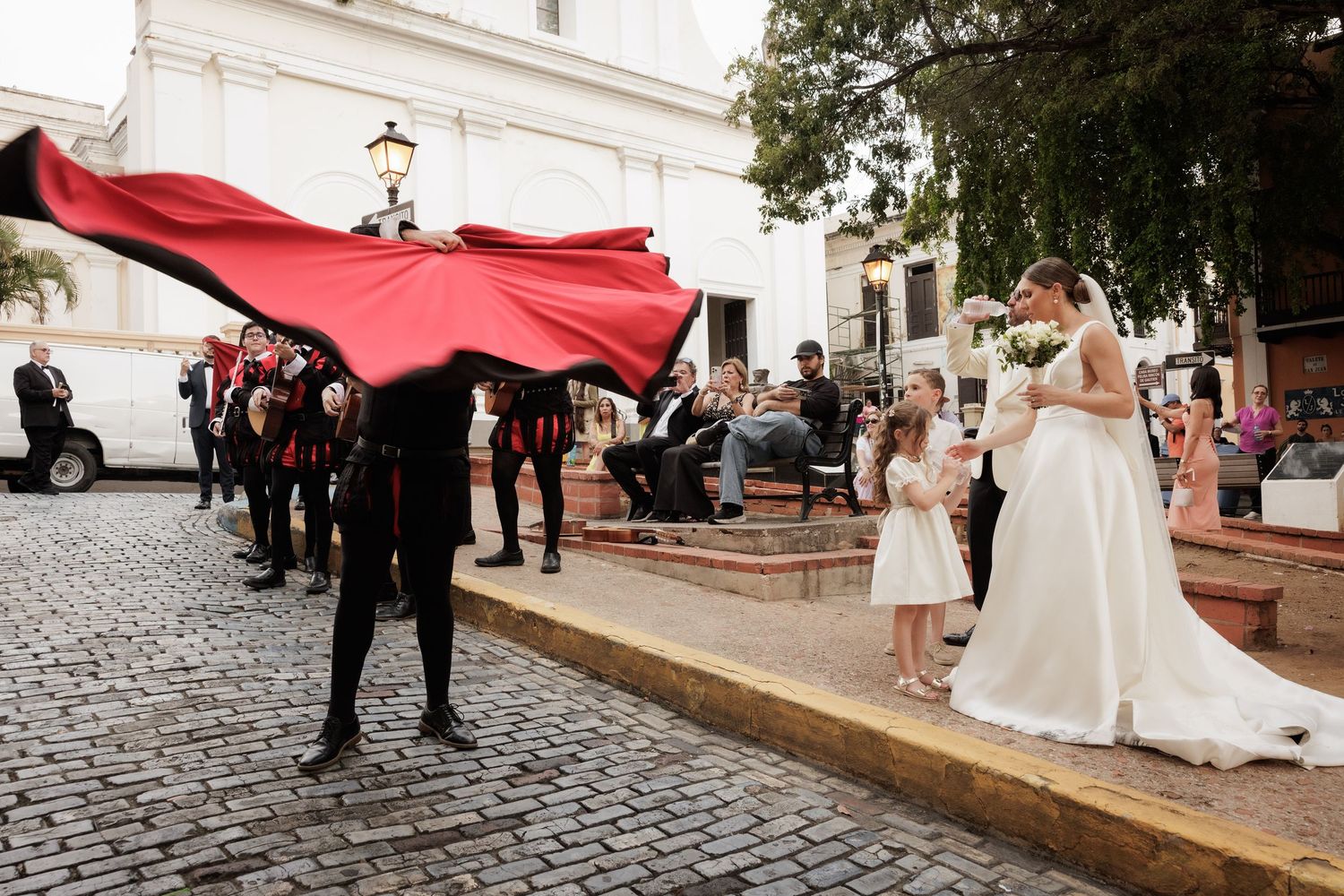 A bride in white dress poses with wedding party members holding a billowing red fabric on a historic street.