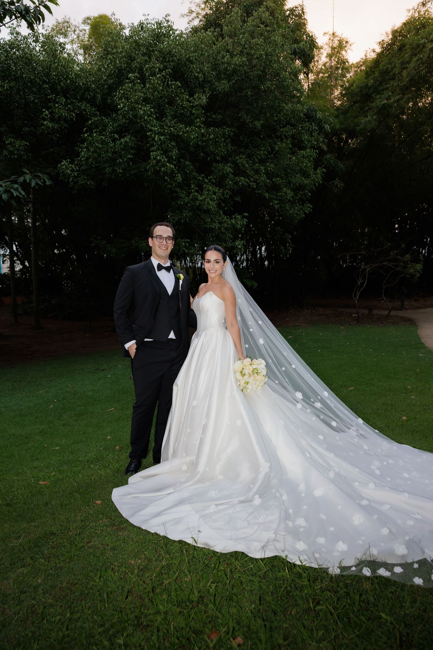 A bride and groom pose together on a grassy lawn at sunset, with her flowing white cathedral veil trailing behind them.