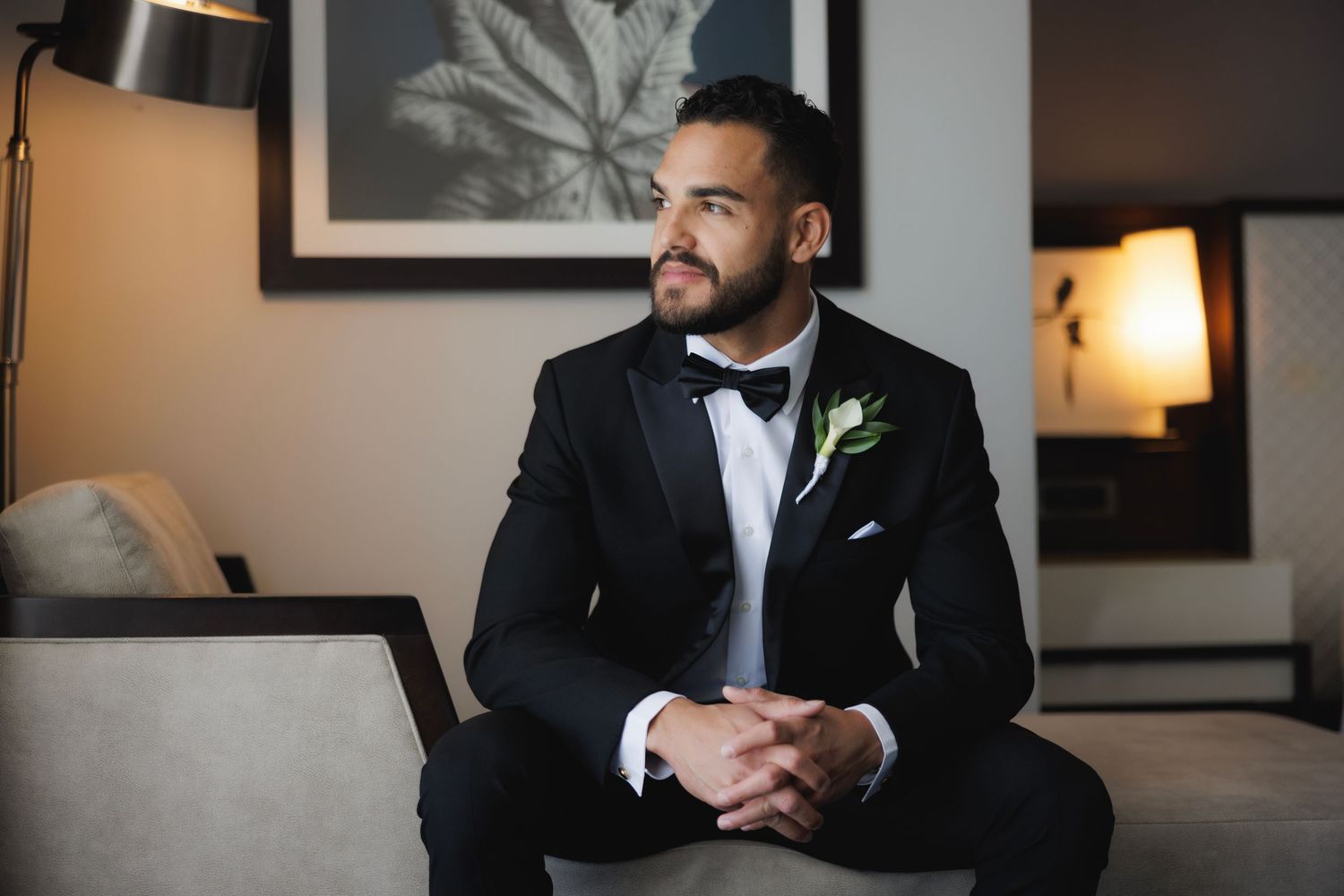 Groom in black tuxedo and bowtie stands contemplatively in elegant hotel room setting.