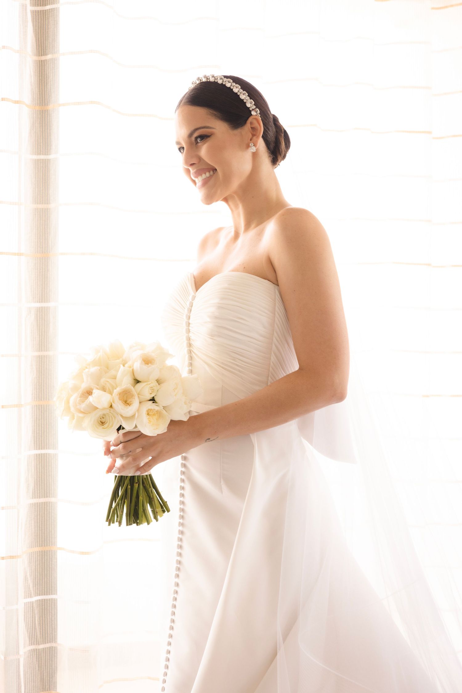 A bride in a classic white wedding dress holds an elegant bouquet of white flowers near sunlit windows.