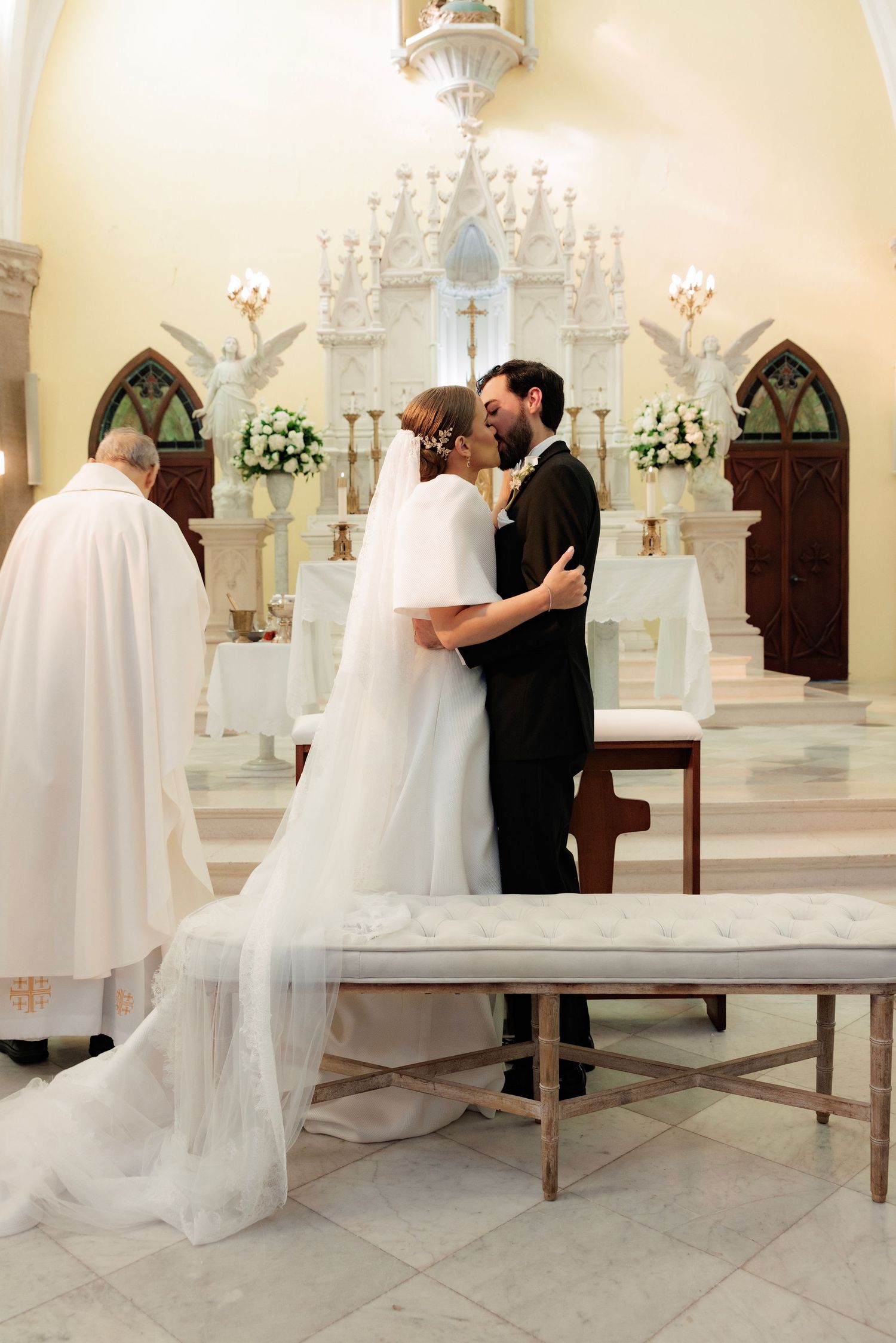 A tender moment during a wedding ceremony in front of an ornate white altar with chandeliers.
