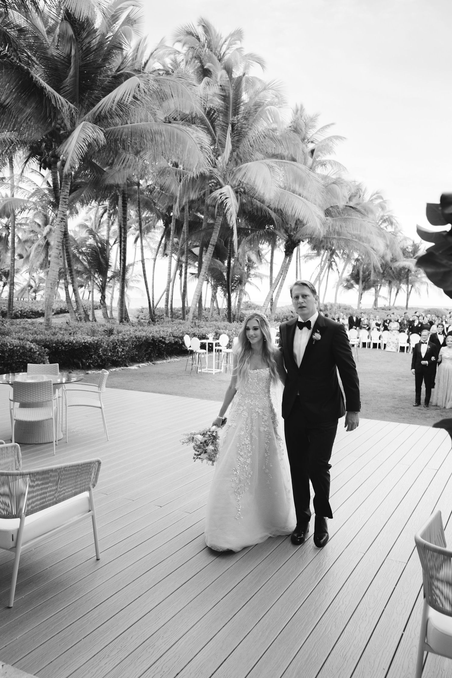 A black and white wedding photo showing a couple walking on a deck surrounded by tropical palm trees.