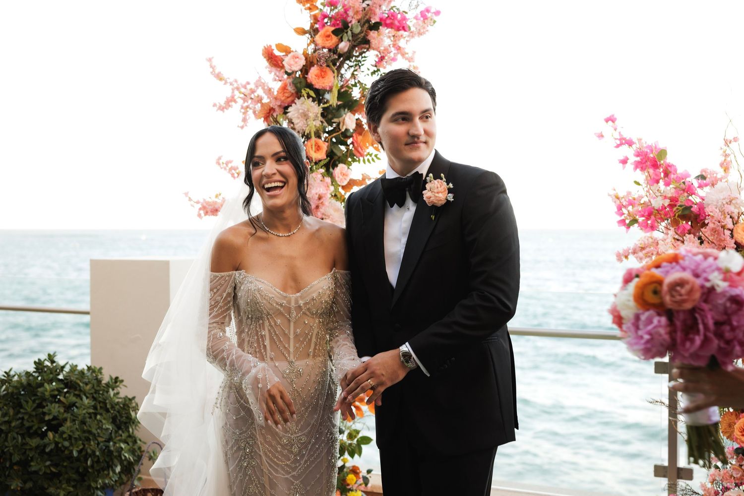 A wedding ceremony by the ocean with pink floral arrangements and formal attire creates an elegant seaside atmosphere.
