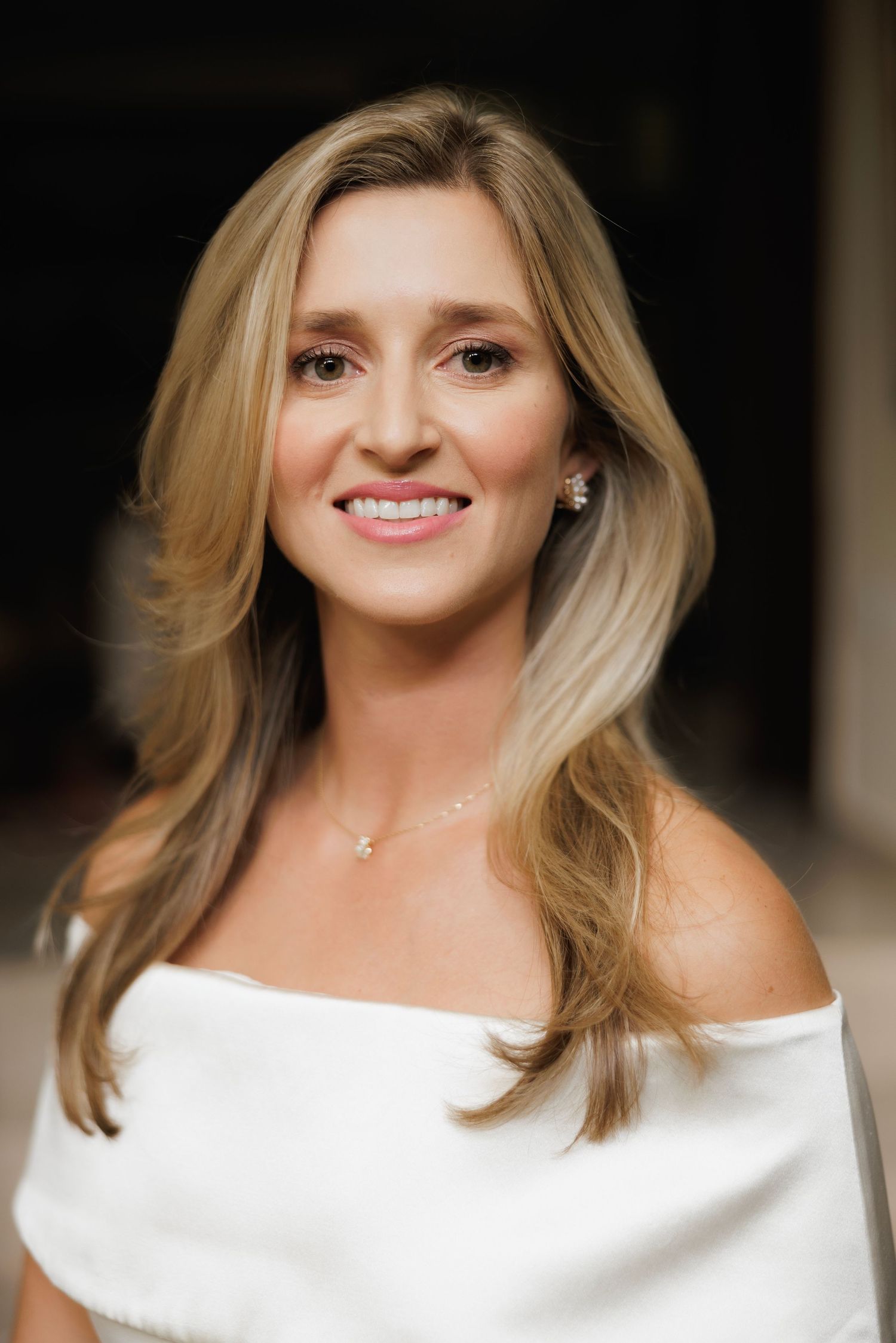 Portrait of a smiling woman with blonde hair wearing an elegant white off-shoulder top against a dark background.
